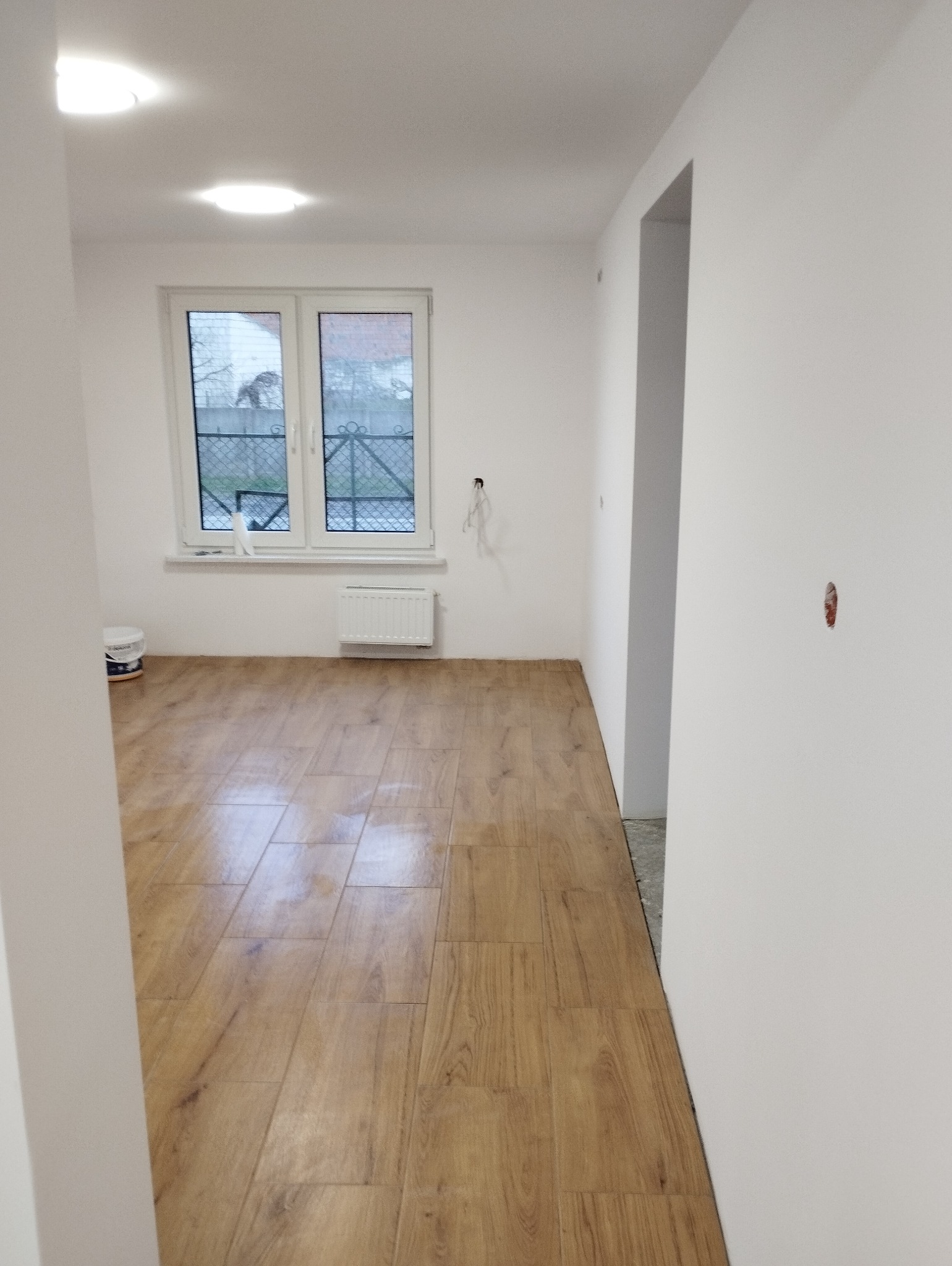 Jasne, wykończone wnętrze z drewnopodobną podłogą, białymi ścianami i oknem. Widoczny fragment grzejnika i puszka farby. Minimalistyczny design, świeżo po remoncie.