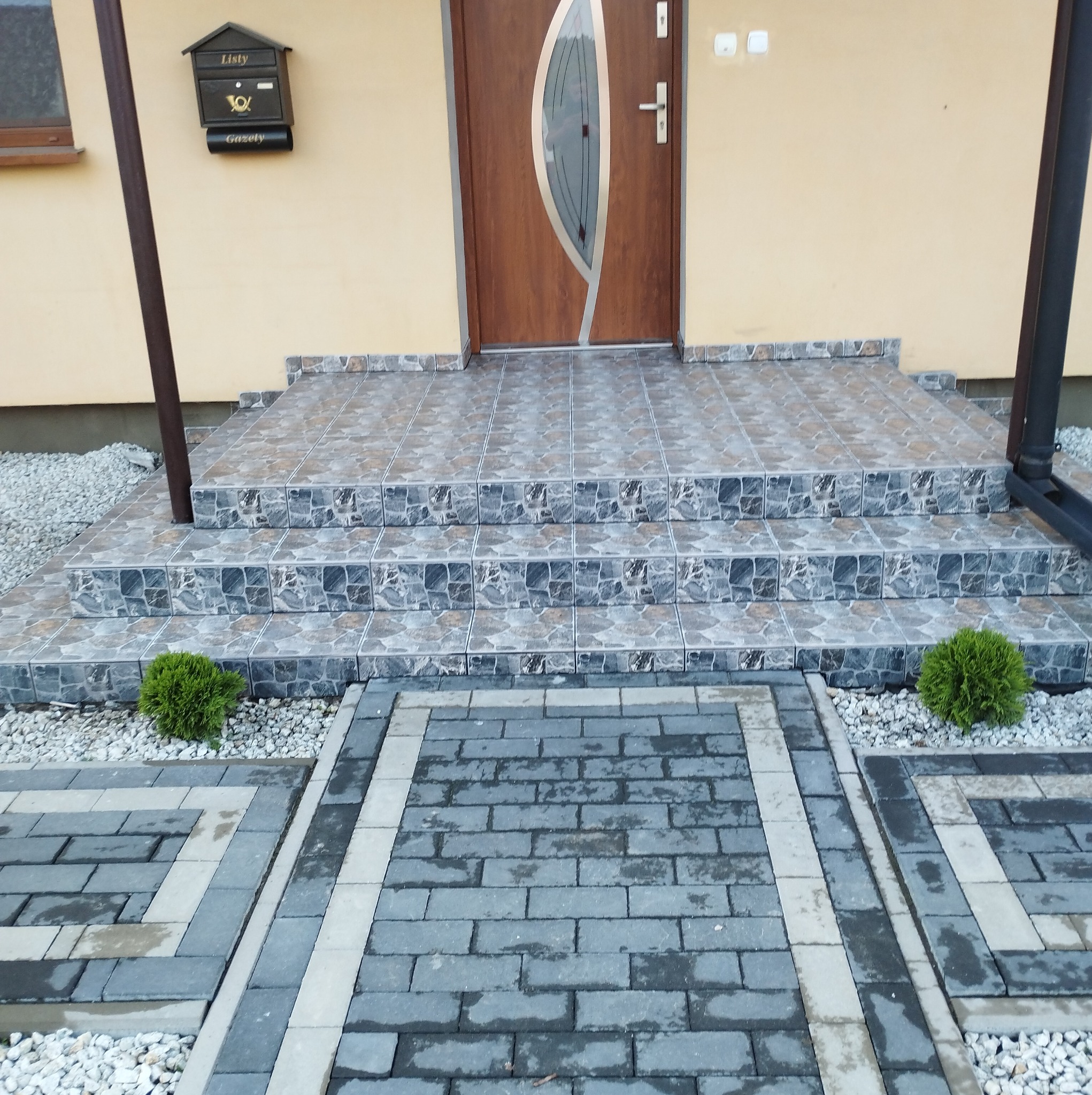 Wykończone płytkami schody wejściowe do domu z brązowymi drzwiami i skrzynką na listy, otoczone szarą kostką brukową i białym żwirem.