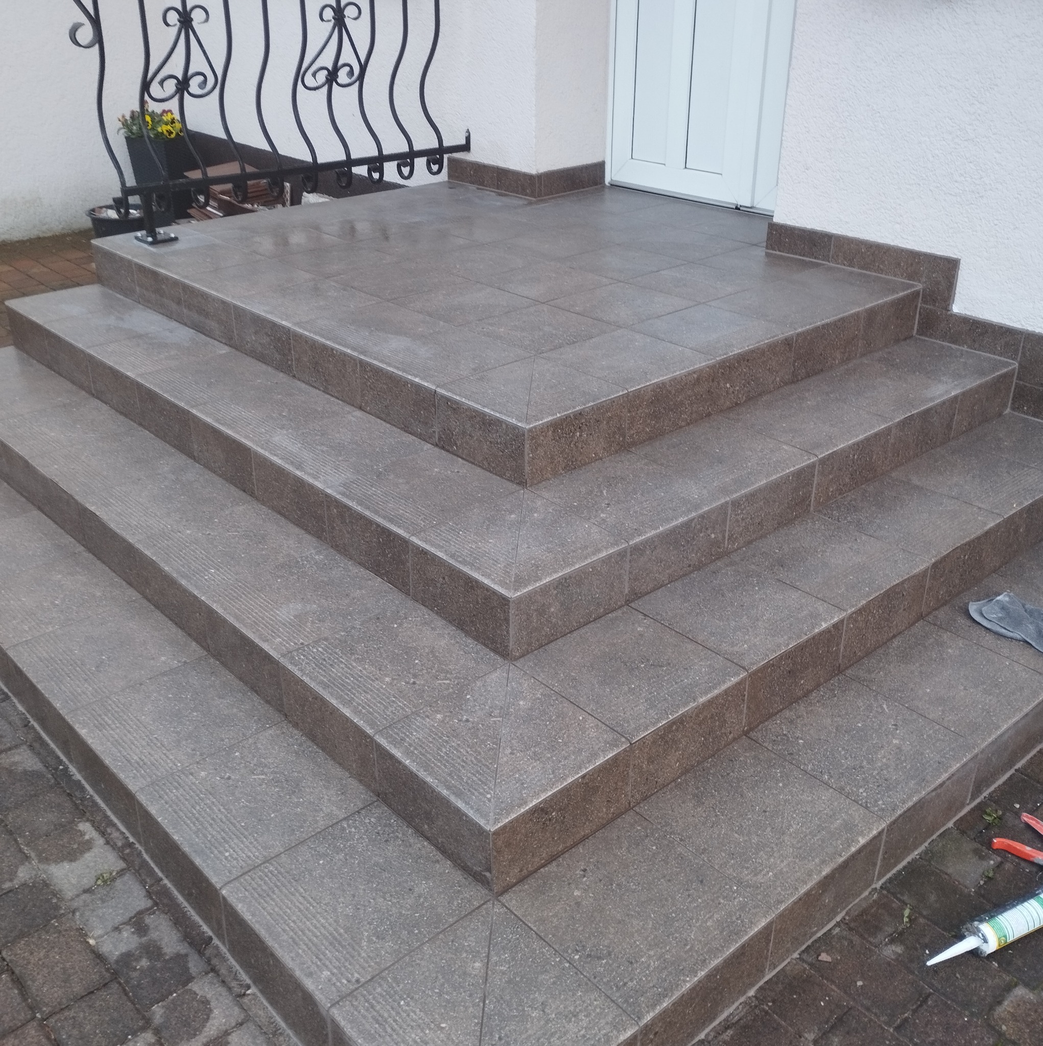 Wykończone płytkami schody wejściowe do domu, z widocznymi narzędziami glazurniczymi. Ujęcie z dołu, podkreślające precyzję ułożenia płytek i estetykę wykończenia.