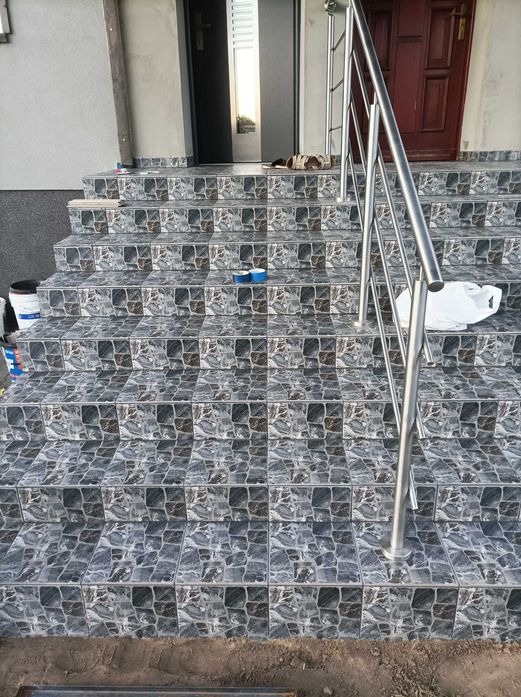 Wykończone płytkami schody wejściowe z metalową barierką. Widoczne detale wzoru płytek i elementy wykończeniowe. Na schodach leżą akcesoria glazurnicze.