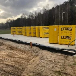 Żółte paczki bloczków YTONG ułożone na betonowym fundamencie, widoczne ślady opon na piasku, w tle drzewa i pochmurne niebo.