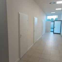 Korytarz biurowy po malowaniu ścian na jasny odcień, z rzędem białych drzwi i jasną podłogą z płytek. Widoczne detale wykończenia i oświetlenie sufitowe.