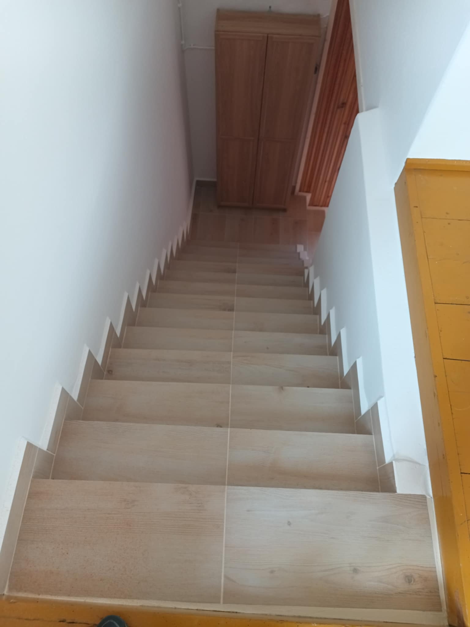 Wykończone schody z jasnymi płytkami imitującymi drewno, białe ściany i żółta framuga drzwi. Widok z góry na dół, z szafką w tle.