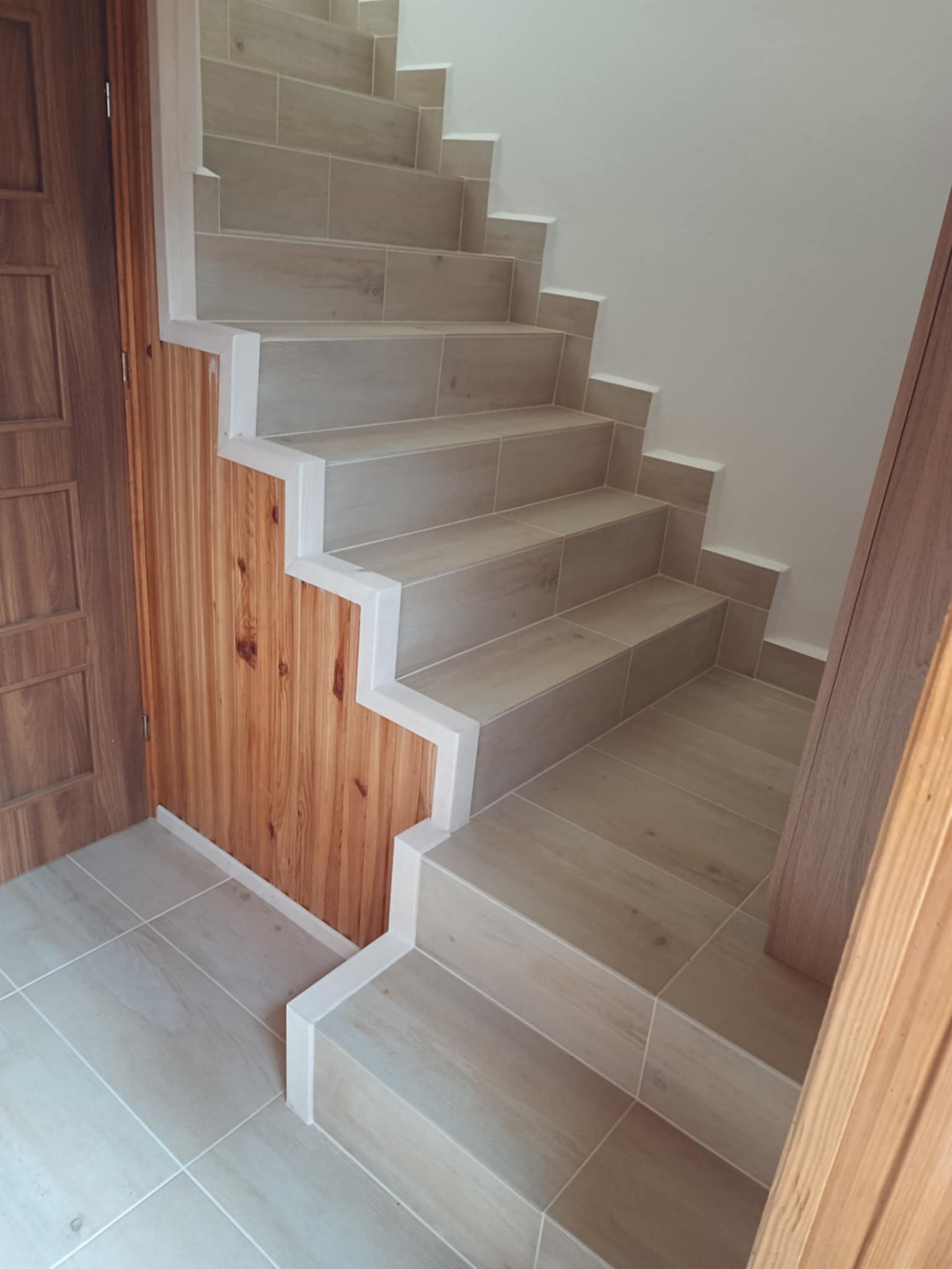 Wykończone schody z płytek w odcieniach beżu, z drewnianą boazerią na ścianie i drewnianymi drzwiami. Widok z dołu, podkreślający detale wykończenia.