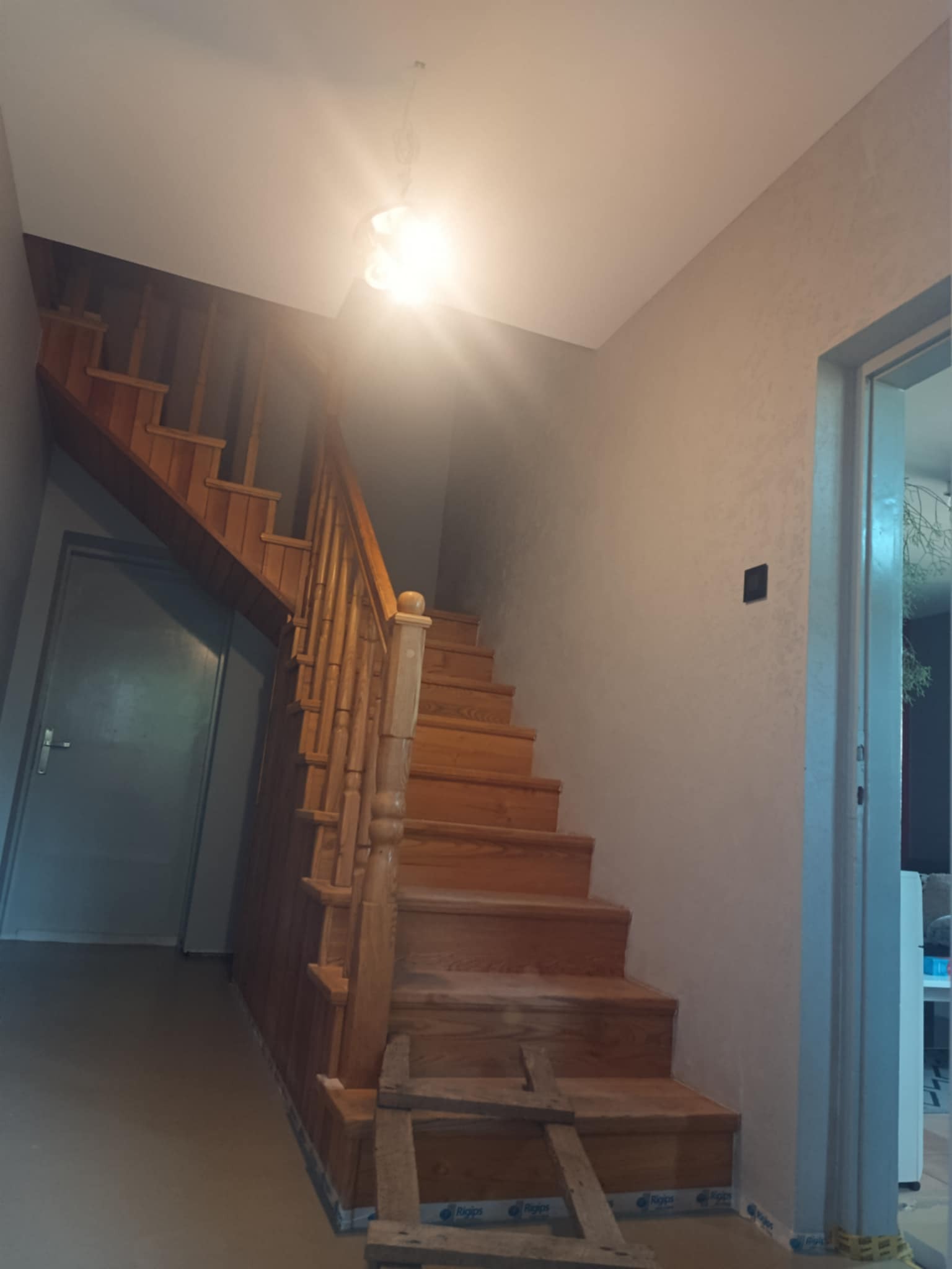 Drewniane schody z balustradą w jasnym wnętrzu po remoncie. Widoczne zabezpieczenia podłogi i ściany, co sugeruje świeżo zakończone prace wykończeniowe.