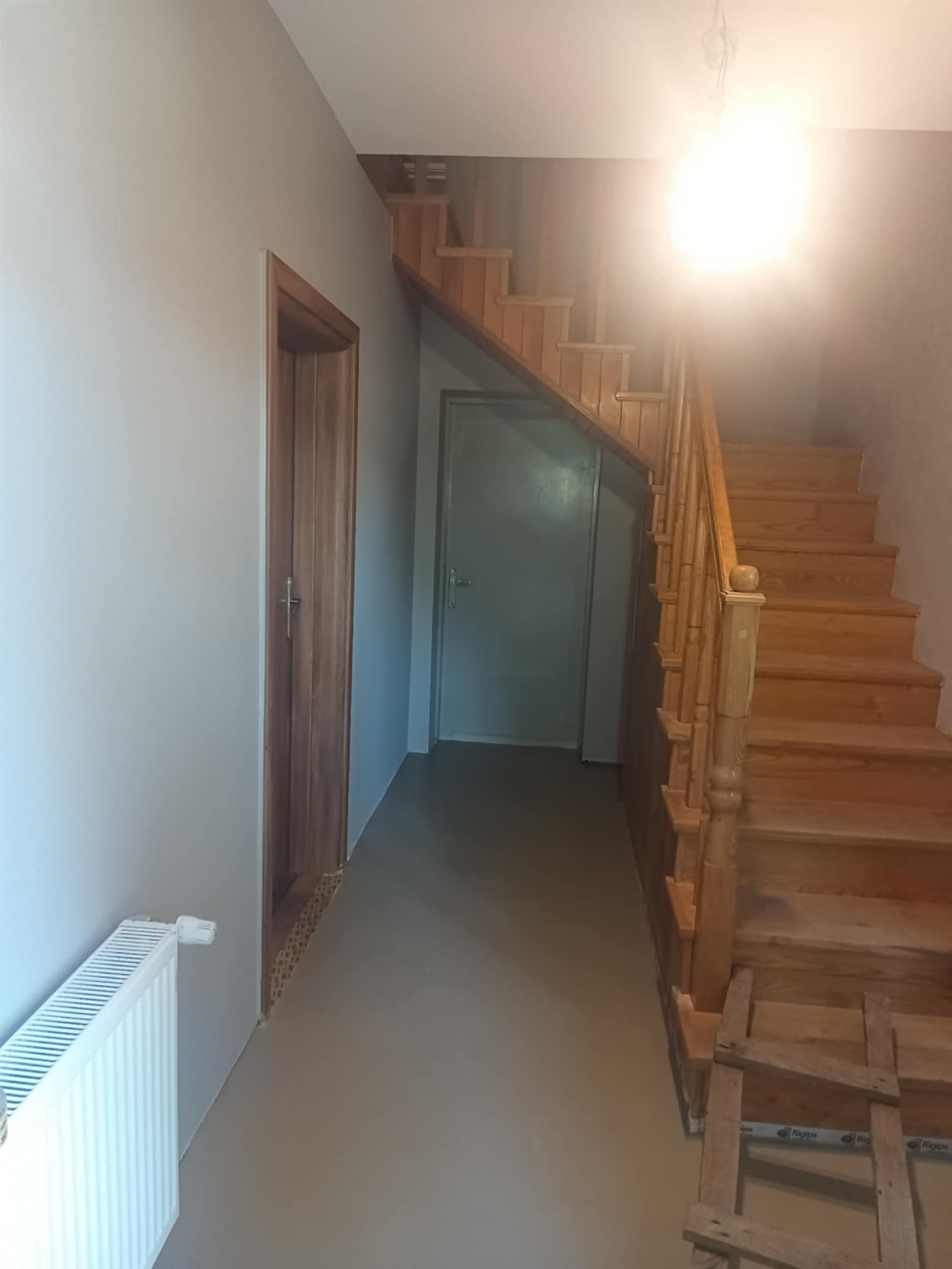 Korytarz po odświeżeniu: szare ściany, drewniane schody i drzwi. Widoczny grzejnik i drabina malarska. Jasne oświetlenie sufitowe.