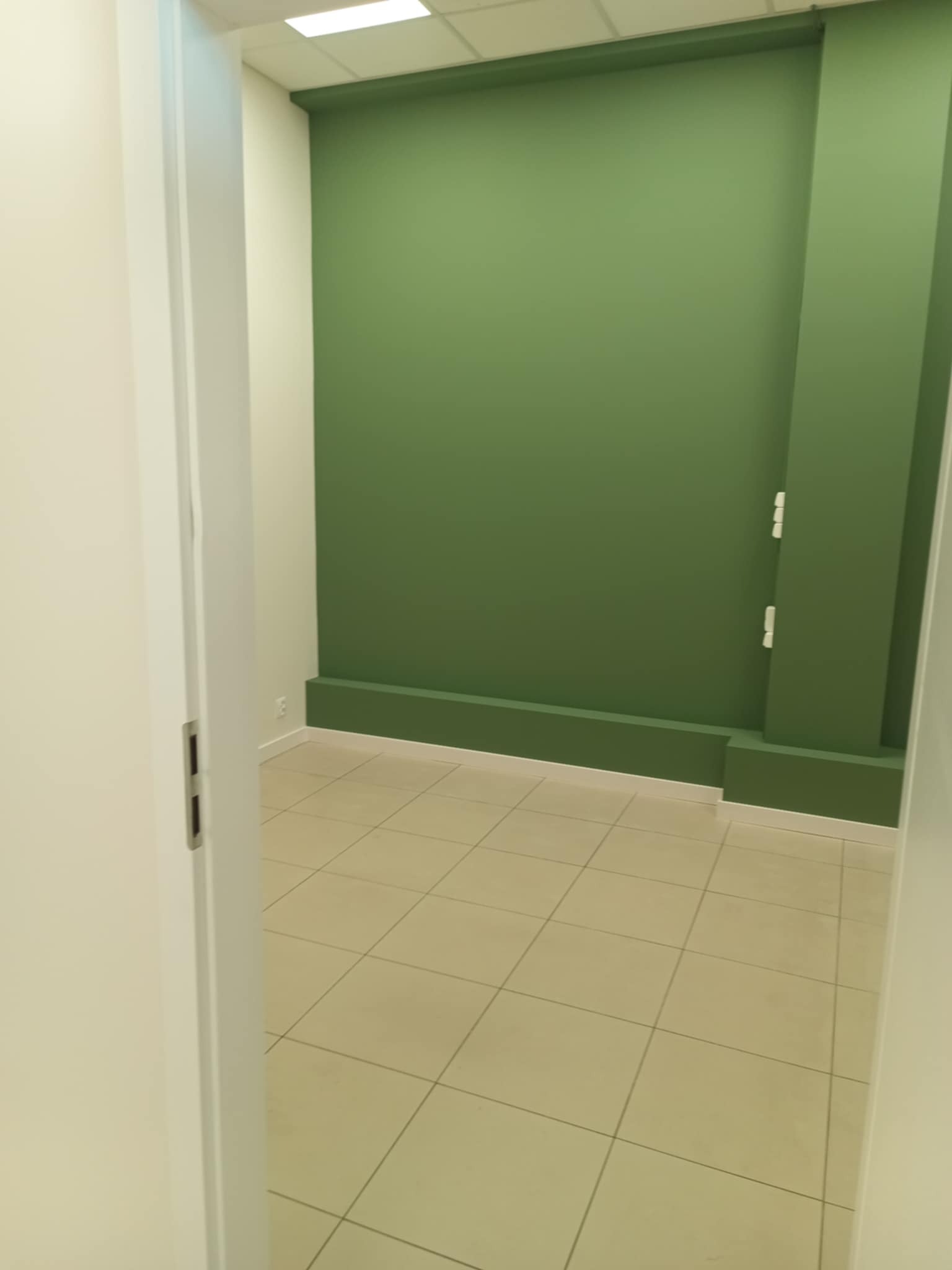 Pomalowane na zielono wnętrze z beżową podłogą z płytek, widoczne przez uchylone białe drzwi. Minimalistyczny design, świeżo po malowaniu.