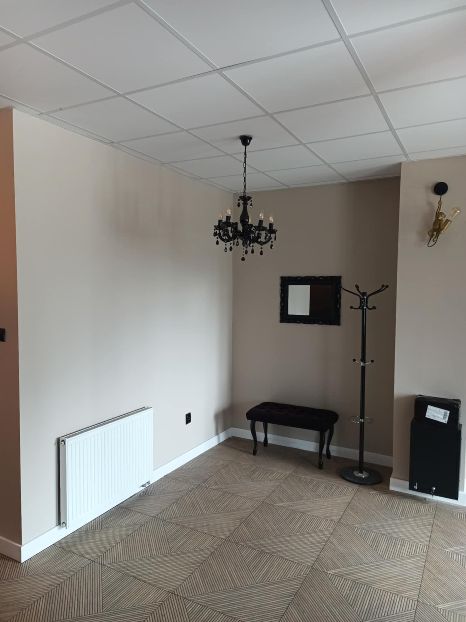 Beżowe wnętrze z grzejnikiem, wzorzystą podłogą, czarnym żyrandolem i wieszakiem. Minimalistyczny wystrój z elementami dekoracyjnymi.