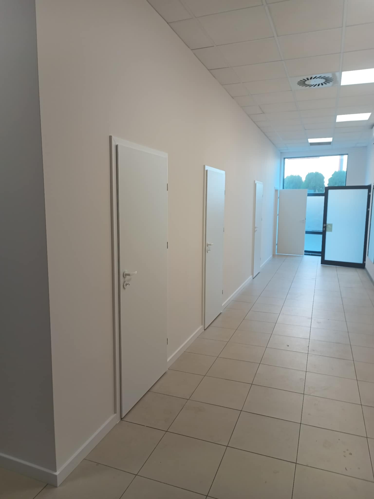 Korytarz biurowy po malowaniu ścian na jasny odcień, z rzędem białych drzwi i jasną podłogą z płytek. Widoczne detale wykończenia i oświetlenie sufitowe.