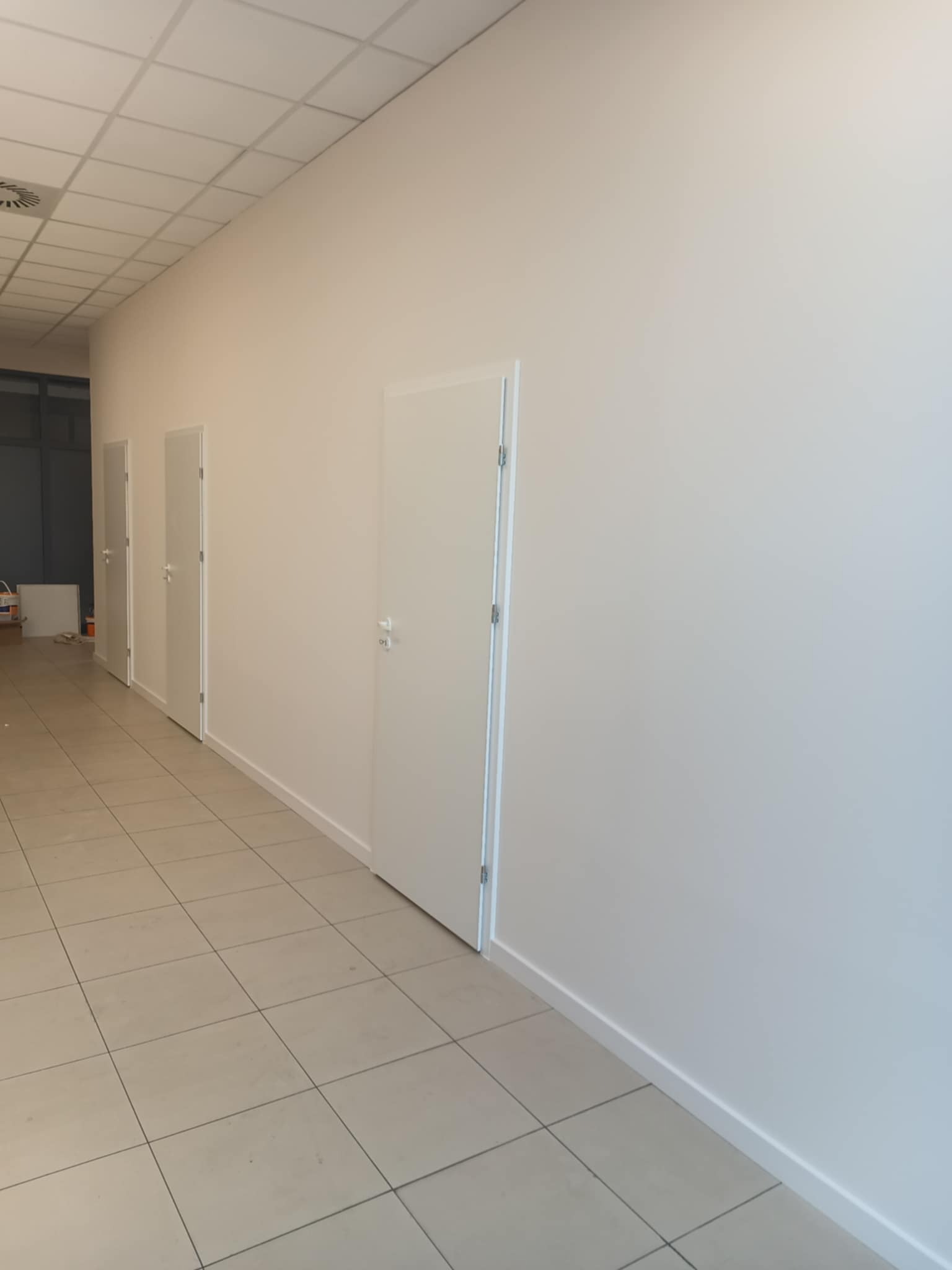 Białe, świeżo pomalowane ściany w korytarzu biurowym z trzema białymi drzwiami. Jasne płytki na podłodze, sufit kasetonowy. Minimalistyczny design, czyste linie.