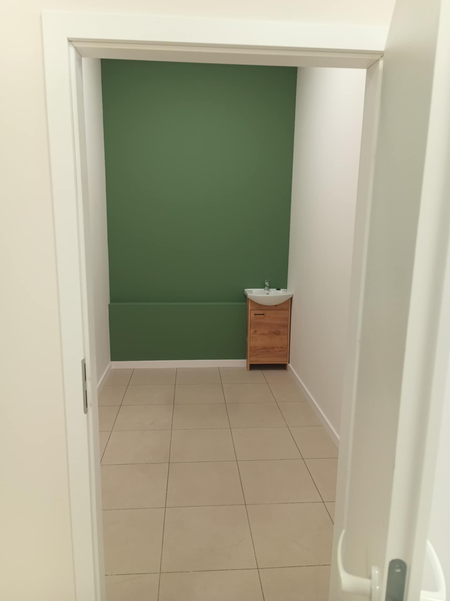 Wykończona łazienka z zieloną ścianą, drewnianą szafką pod umywalką i beżowymi płytkami podłogowymi, widoczna zza otwartych, białych drzwi.