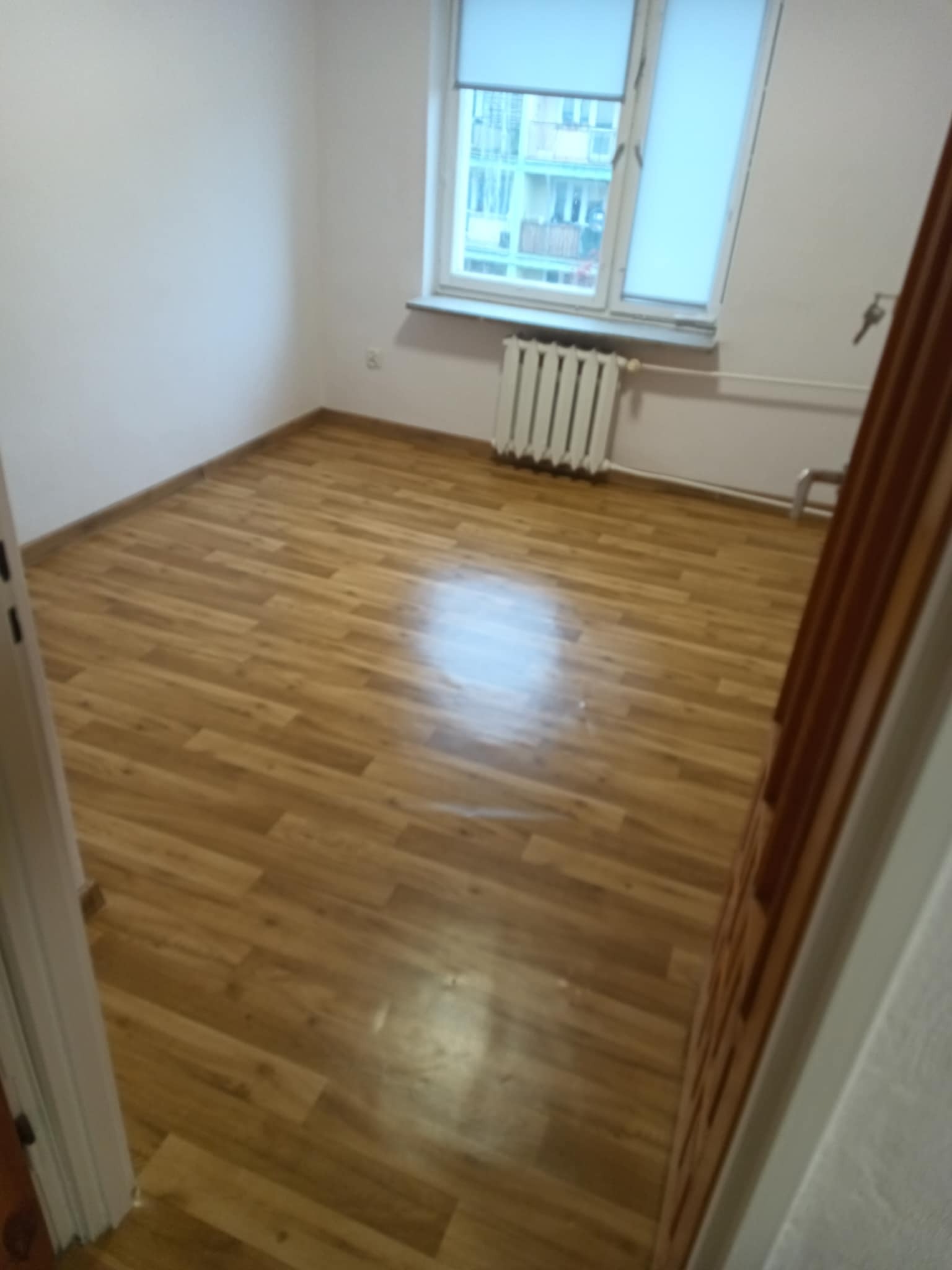 Pokój po remoncie z nową podłogą z paneli imitujących drewno. Widoczne okno z grzejnikiem i fragment drzwi. Ściany pomalowane na biało.