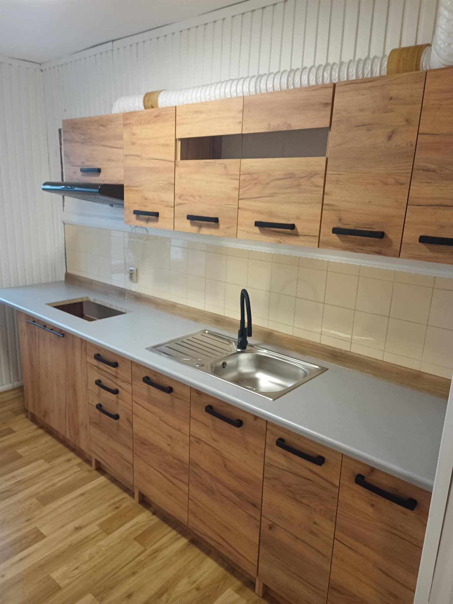 Nowoczesna kuchnia z drewnianymi szafkami i szarym blatem, widok na zlew i baterię. Białe panele ścienne i wentylacja nad szafkami dodają charakteru.