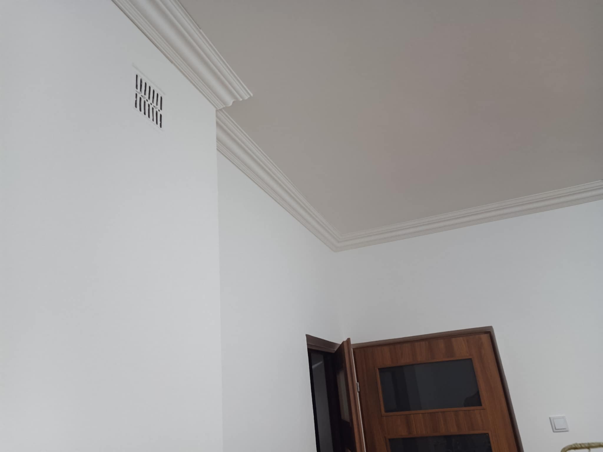 Białe ściany z dekoracyjną listwą przysufitową i kratką wentylacyjną. Widoczny fragment drewnianych drzwi. Minimalistyczne wykończenie wnętrza.
