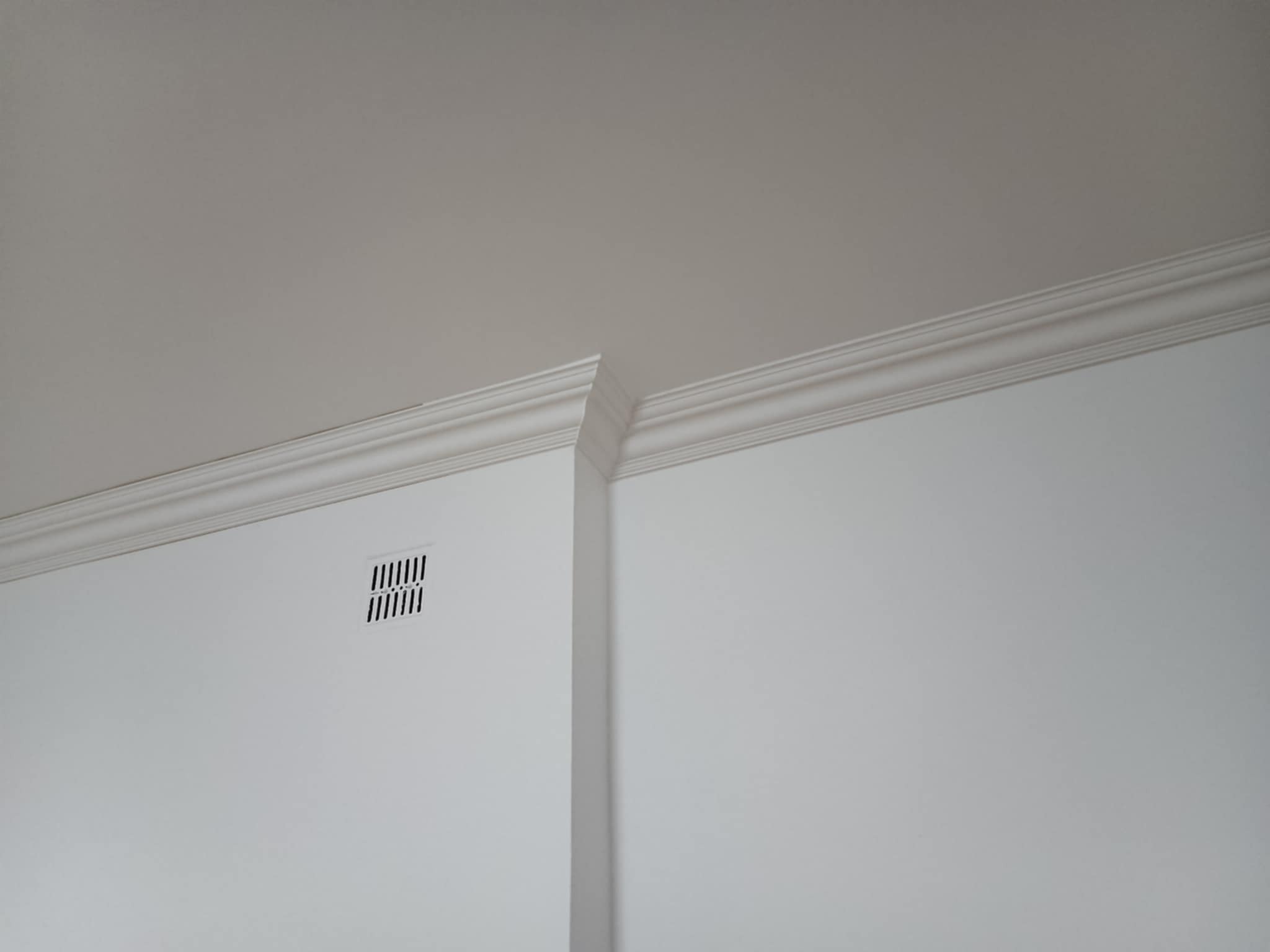 Białe ściany z listwami dekoracyjnymi przy suficie i pionową listwą narożną. Widoczna kratka wentylacyjna. Minimalistyczne wykończenie wnętrza.