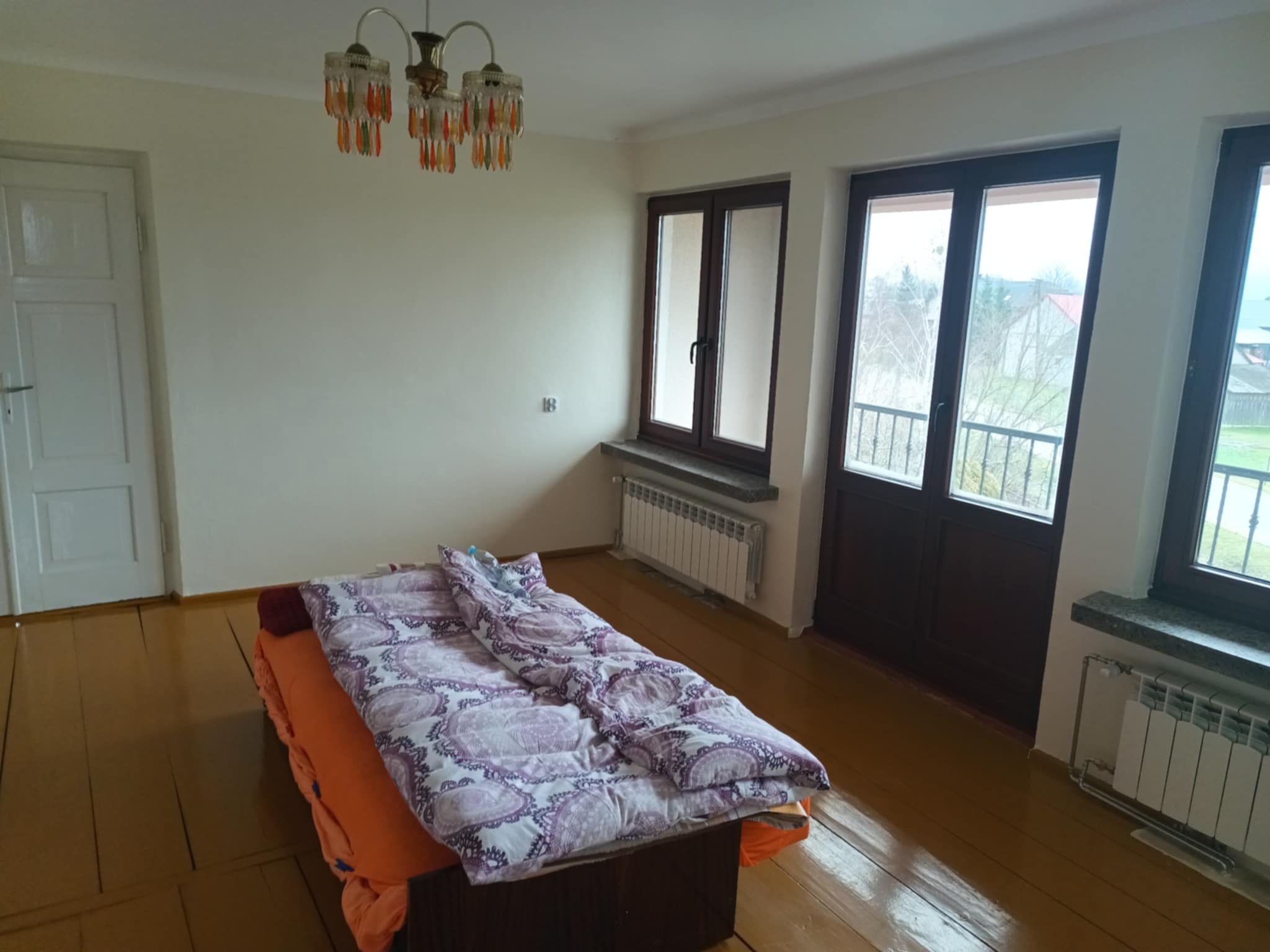 Jasne wnętrze pokoju z drewnianą podłogą, łóżkiem z wzorzystą pościelą i oknami balkonowymi. Widoczny grzejnik pod oknem i ozdobny żyrandol.