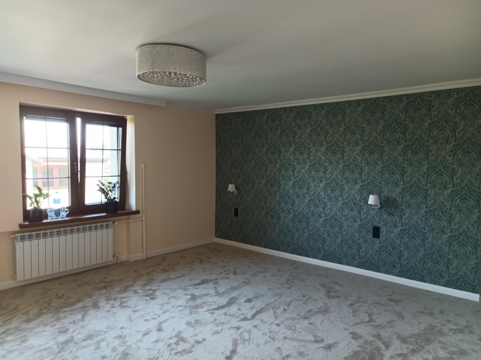 Pokój po remoncie: wzorzysta tapeta na ścianie, jasna podłoga, okno z roślinami i nowoczesna lampa sufitowa. Widok na odnowione wnętrze z dekoracyjnymi detalami.