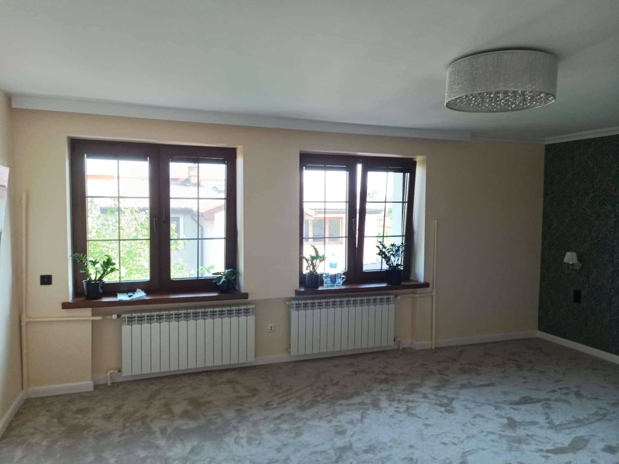 Jasne wnętrze po odświeżeniu: kremowe ściany, ciemne okna z roślinami, białe grzejniki, wzorzysta tapeta na fragmencie ściany i nowoczesna lampa sufitowa.