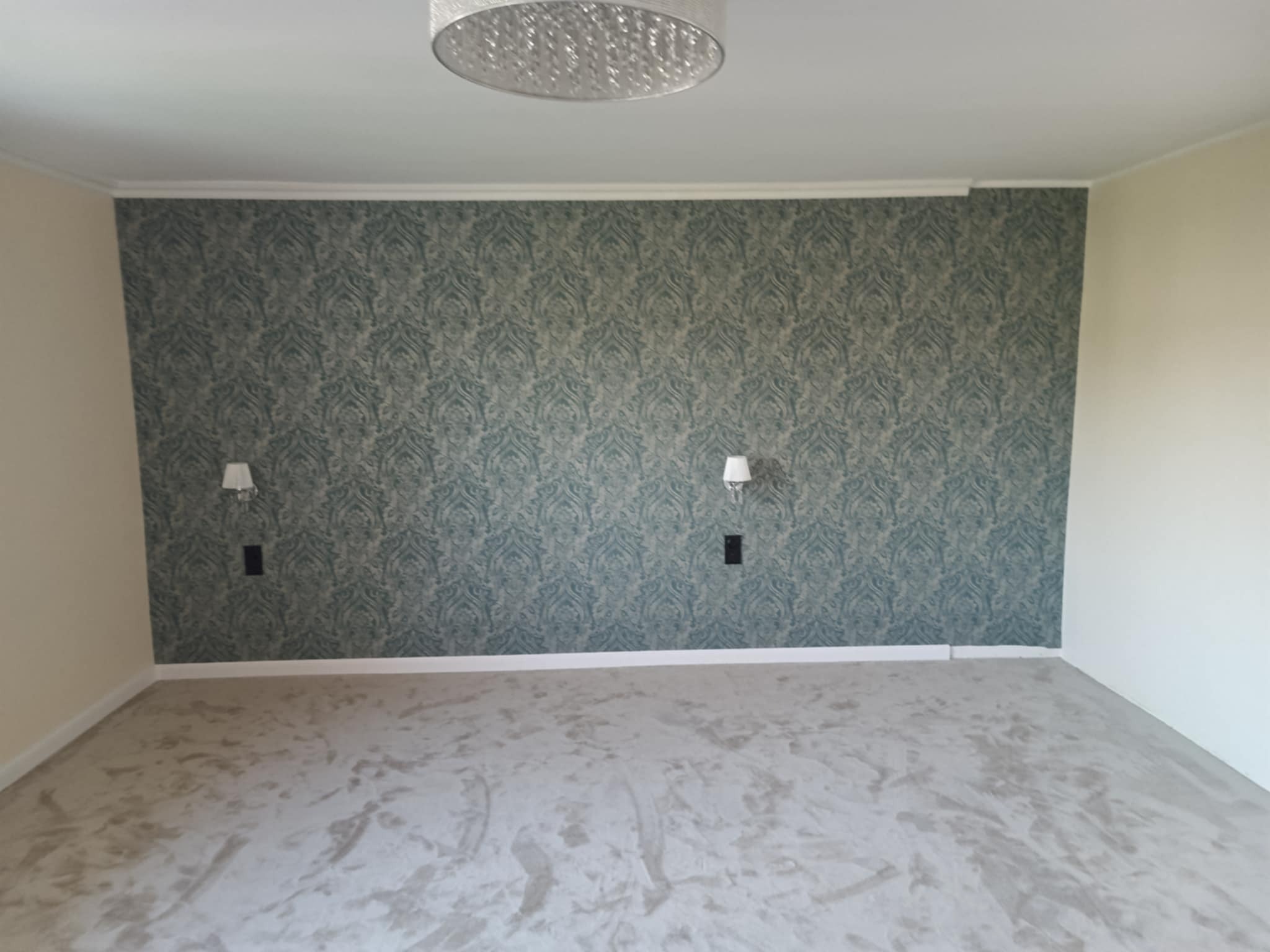 Pokój po tapetowaniu ściany z wzorem w odcieniach zieleni i szarości. Na ścianie zamontowane kinkiety. Na suficie widoczna lampa. Podłoga pokryta wykładziną.