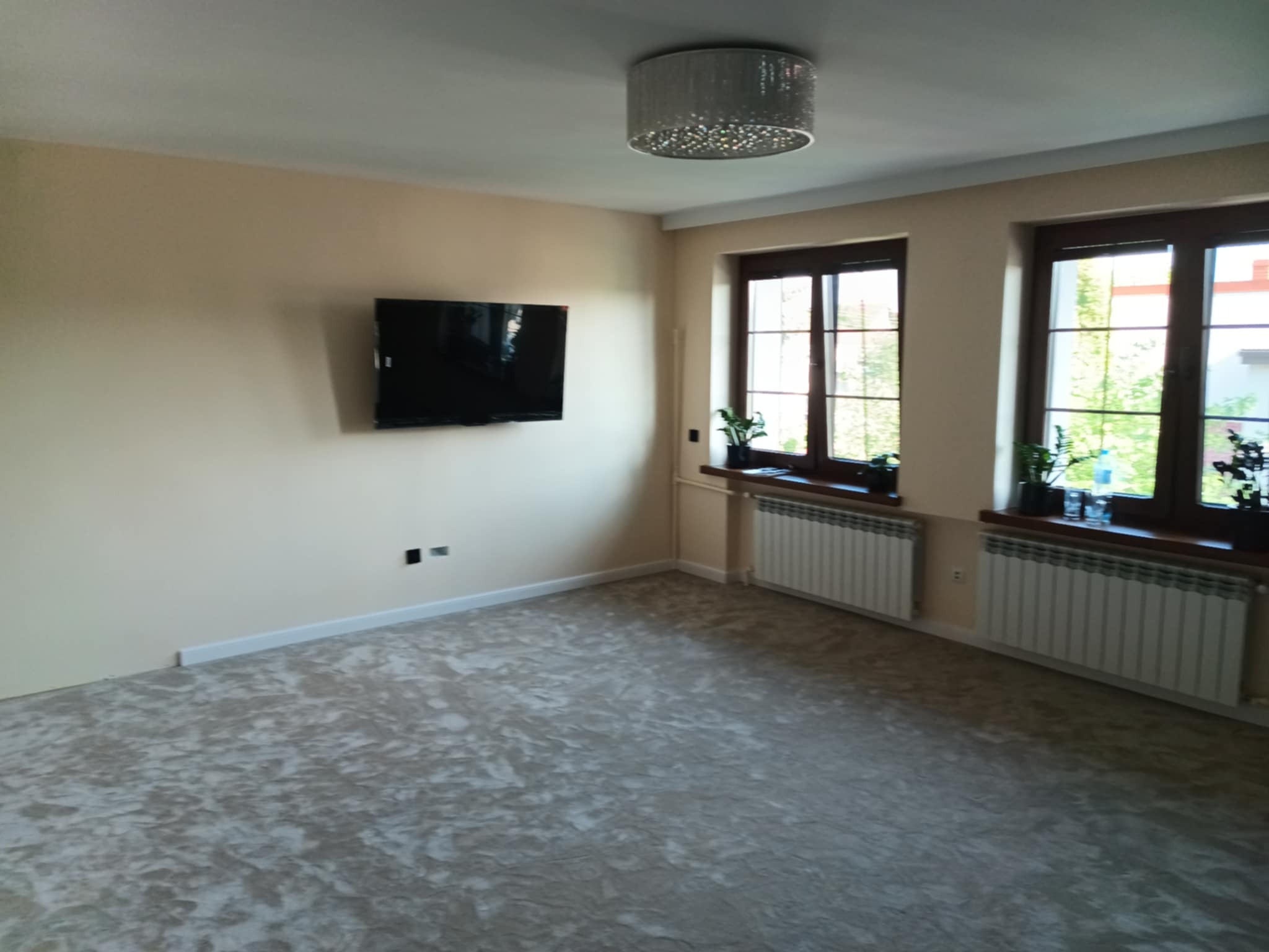 Jasne wnętrze po odświeżeniu: kremowe ściany, dekoracyjna podłoga imitująca kamień, ciemne ramy okien z roślinami i nowoczesny żyrandol. Telewizor na ścianie.