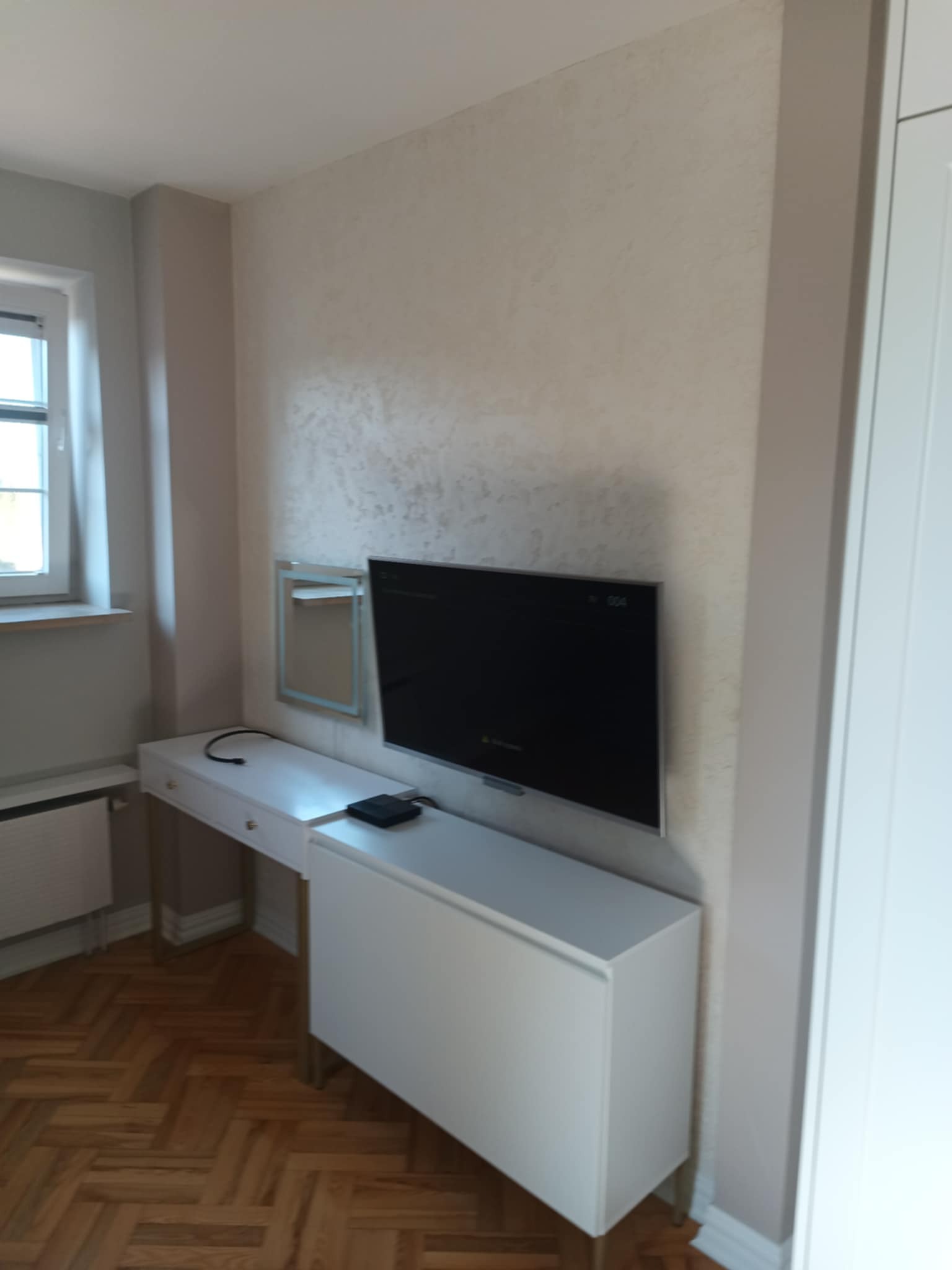 Biała toaletka z lustrem i szafką RTV pod telewizorem na ścianie z efektem dekoracyjnym. Parkiet w jodełkę na podłodze. Nowoczesne wykończenie wnętrza.