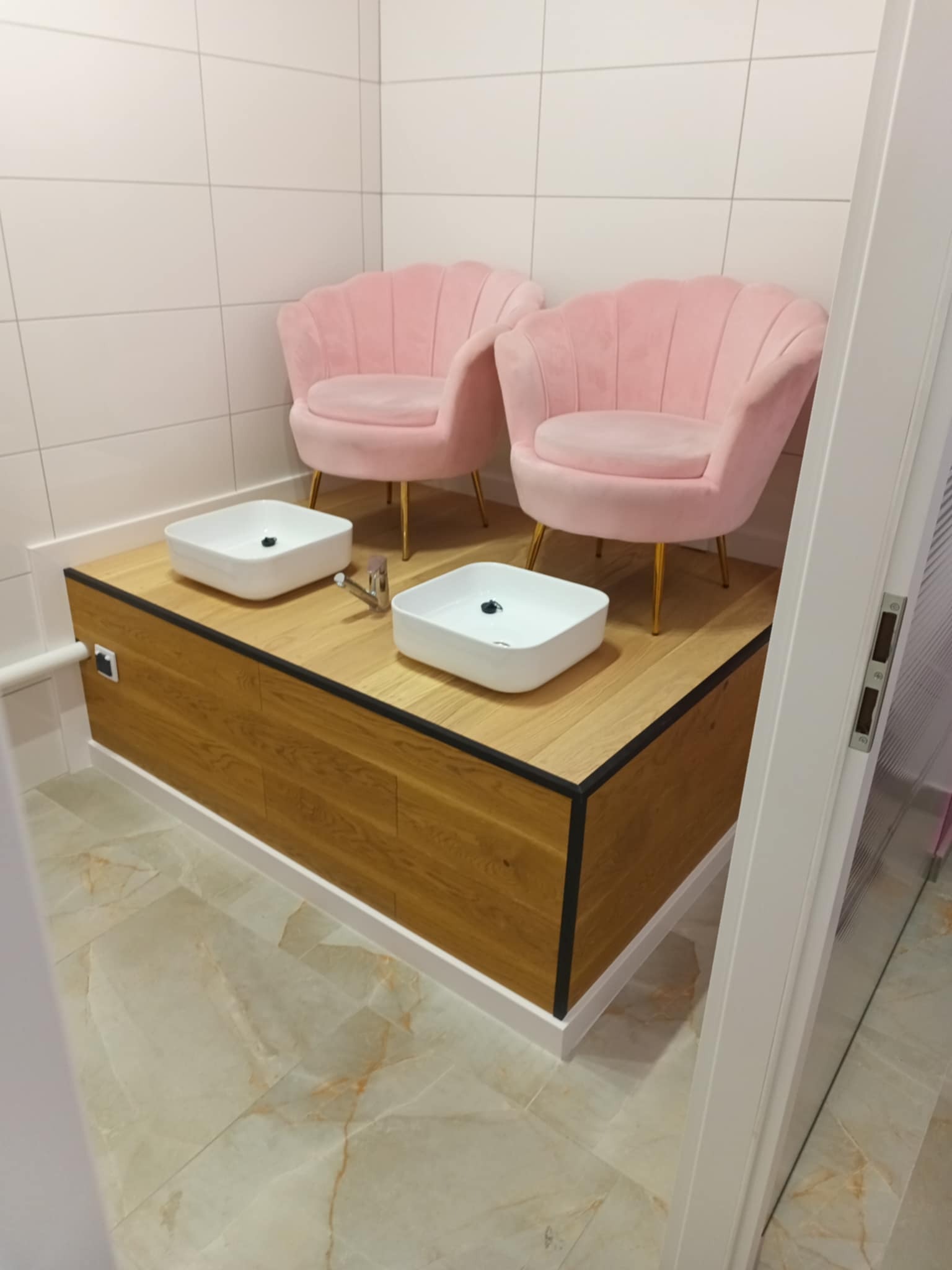 Nowoczesne stanowisko do pedicure z dwoma umywalkami i różowymi fotelami w salonie kosmetycznym. Jasne płytki na ścianach i marmurowa podłoga.