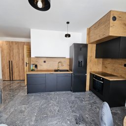 Anna Jaworowska - Nowoczesna kuchnia z czarnymi szafkami, drewnianymi akcentami i marmurową podłogą. Złota bateria i minimalistyczne oświetlenie dodają elegancji. Widok na salon.