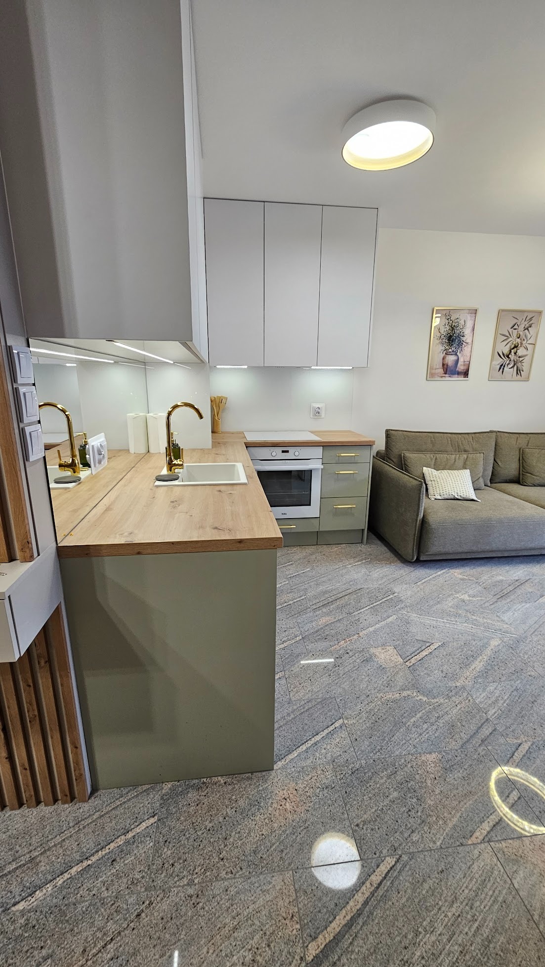 Nowoczesna kuchnia z drewnianym blatem i złotą armaturą, połączona z salonem. Szare płytki podłogowe z subtelnym wzorem. Jasne, minimalistyczne szafki.