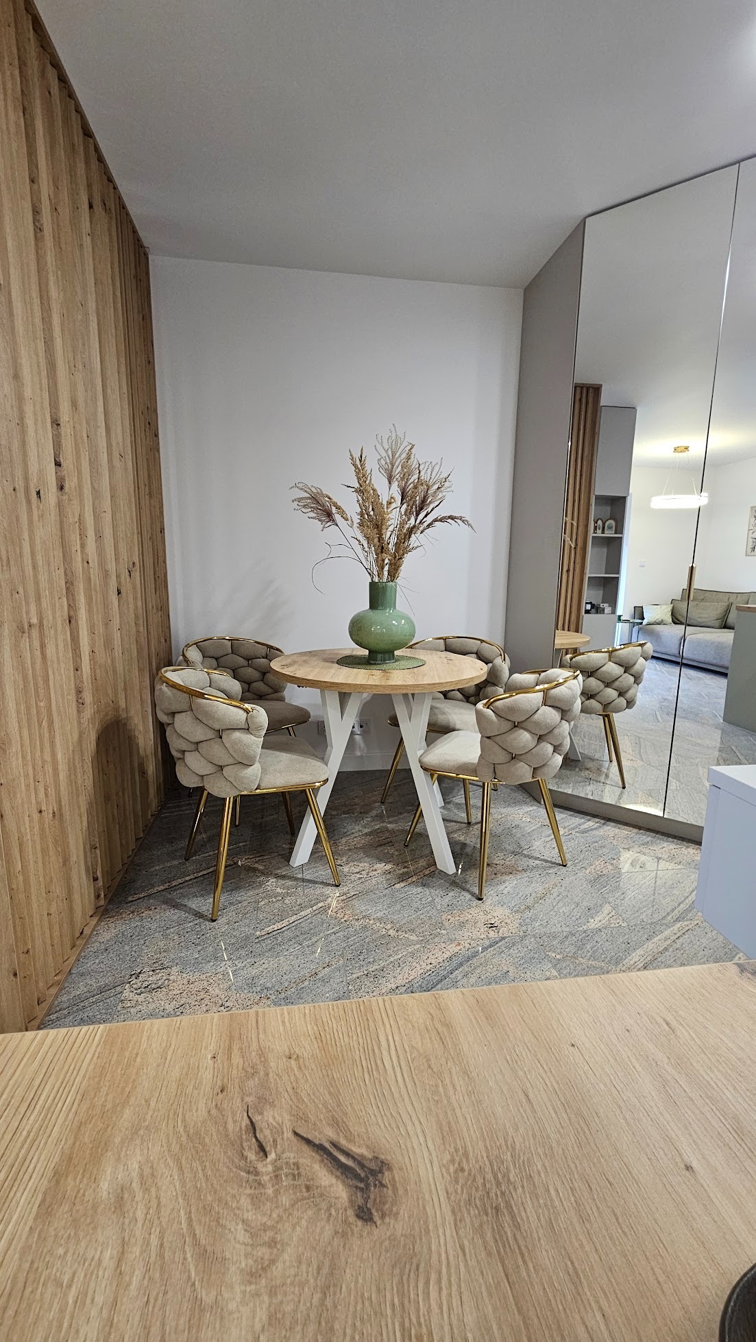 Jadalnia z okrągłym stołem na białych nogach i czterema krzesłami z beżową tapicerką i złotymi nogami. Na stole zielony wazon z suchymi trawami. Lustro w tle.