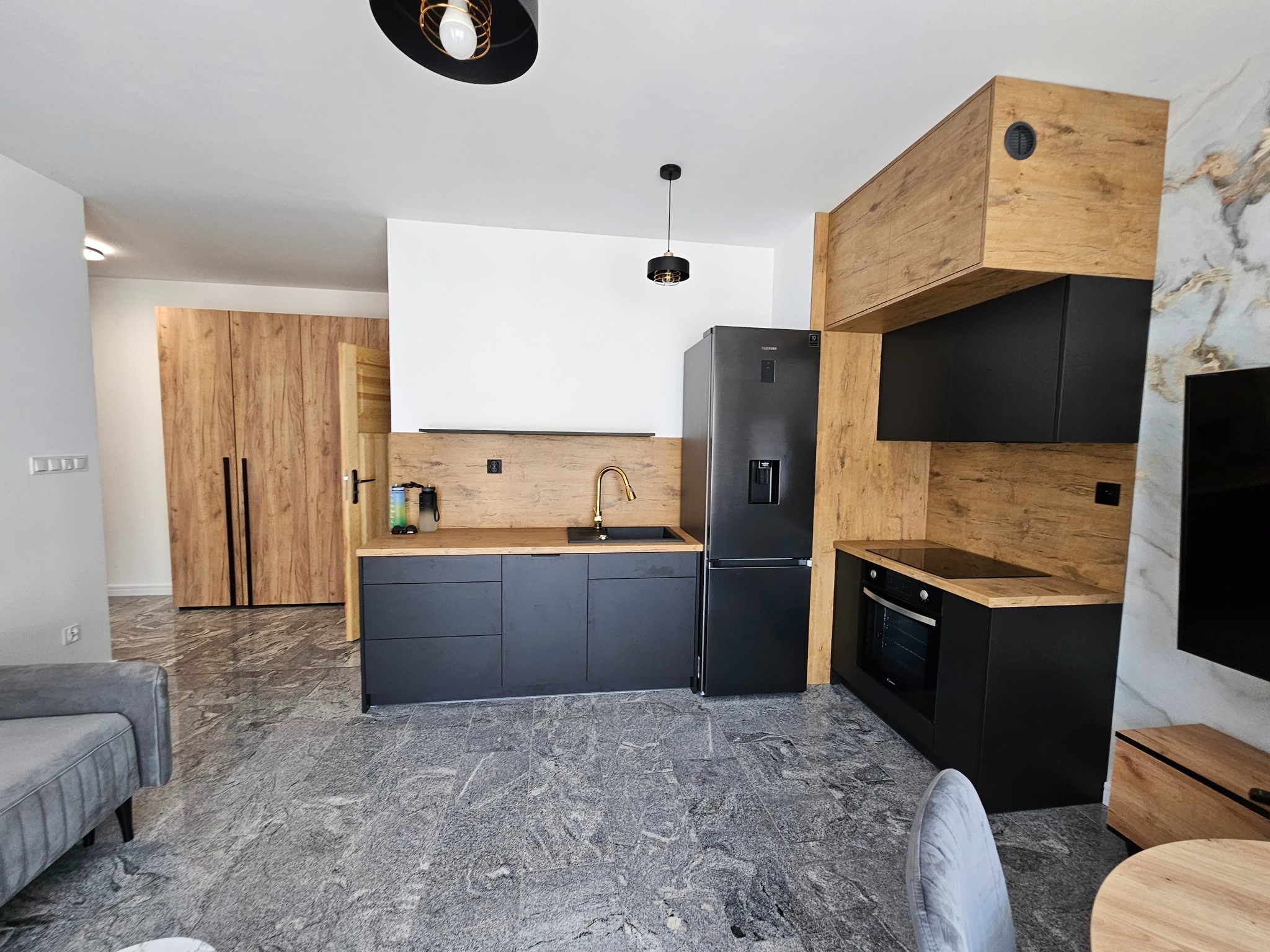 Nowoczesna kuchnia z czarnymi szafkami, drewnianymi akcentami i marmurową podłogą. Złota bateria i minimalistyczne oświetlenie dodają elegancji. Widok na salon.