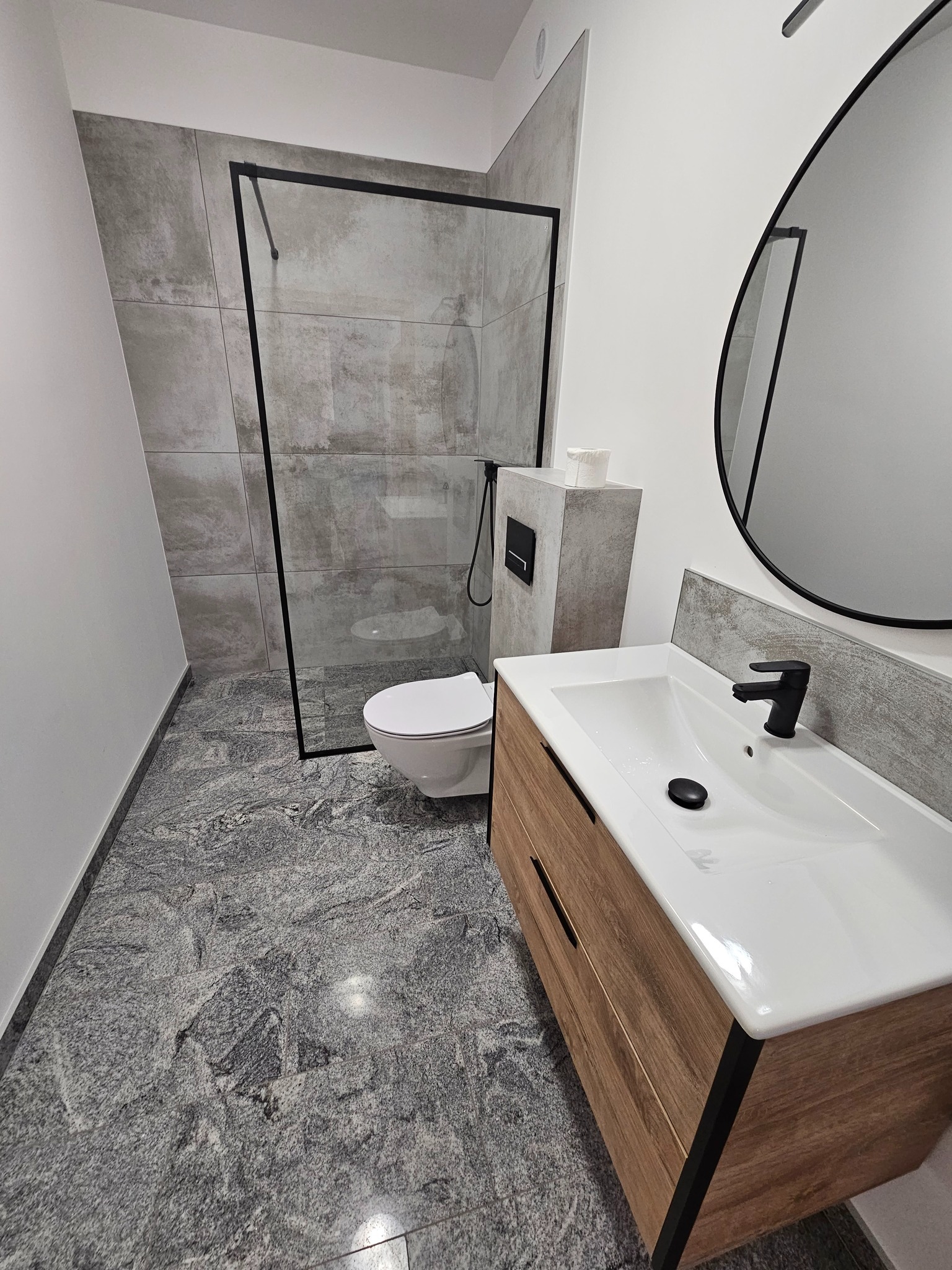 Nowoczesna łazienka z kabiną typu walk-in, umywalką z drewnianą szafką i okrągłym lustrem. Szare płytki na ścianach i podłodze tworzą spójną całość.