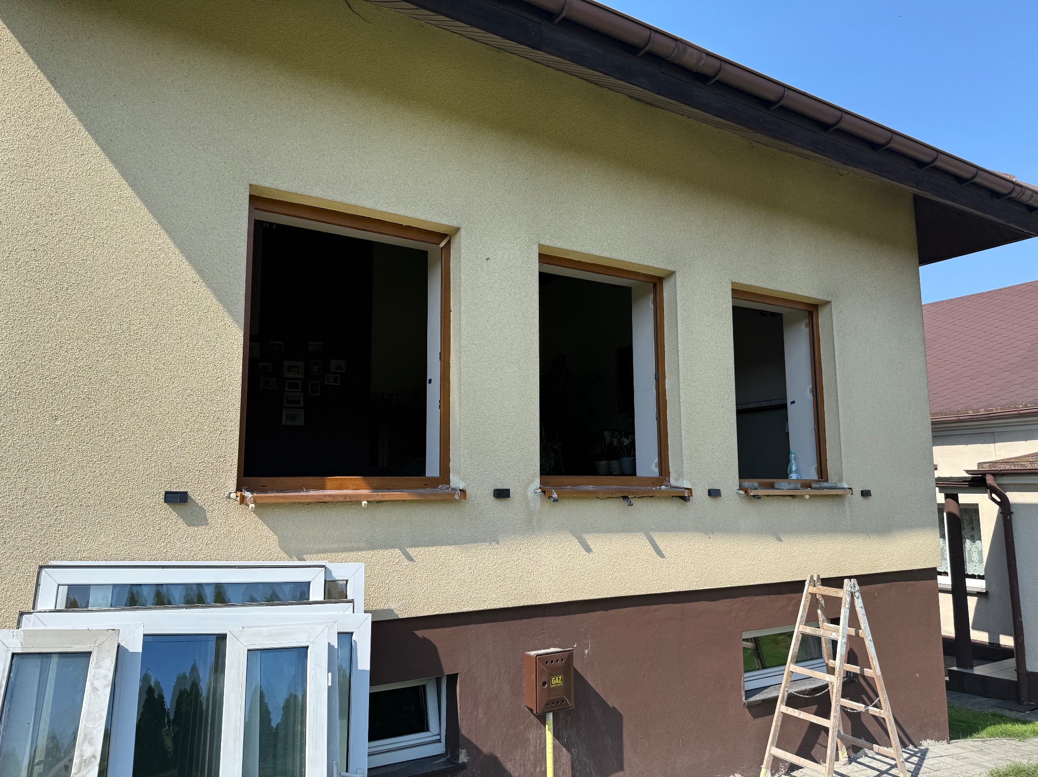Dom z trzema otworami okiennymi w trakcie montażu. Na dole leżą białe ramy okienne, obok stoi drabina. Widoczny fragment elewacji w odcieniach beżu i brązu.