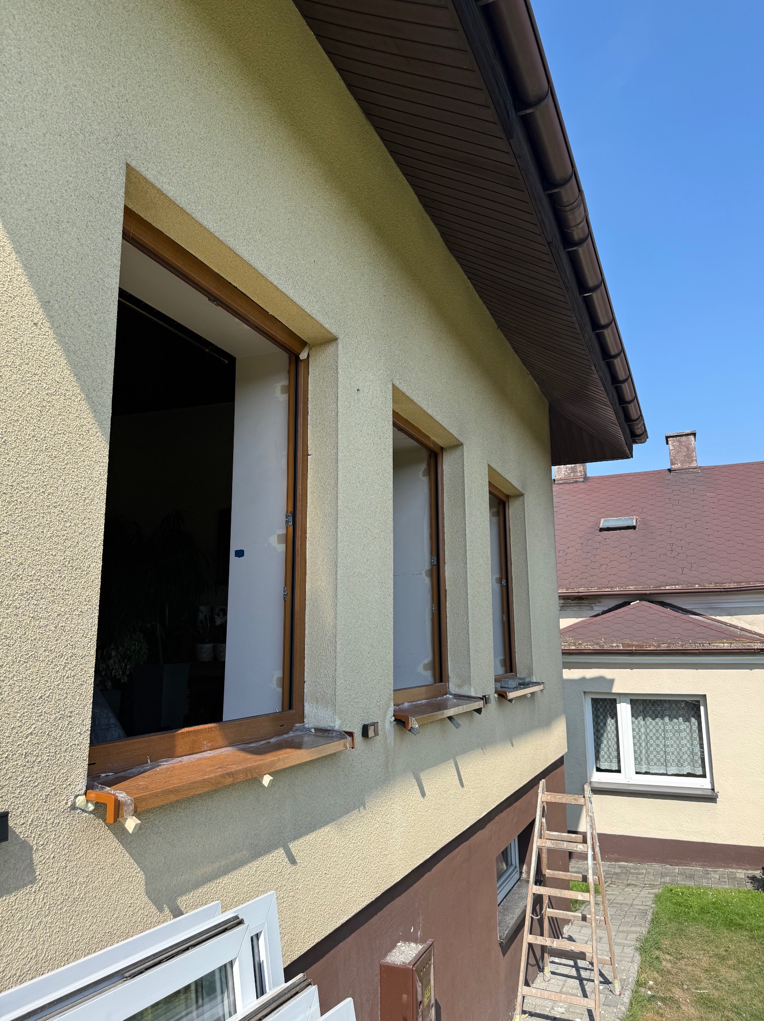 Dom z oknami w trakcie montażu, widoczne drewniane ramy i białe skrzydła okienne. Na dole leżą nowe okna, a obok ściany stoi drabina.