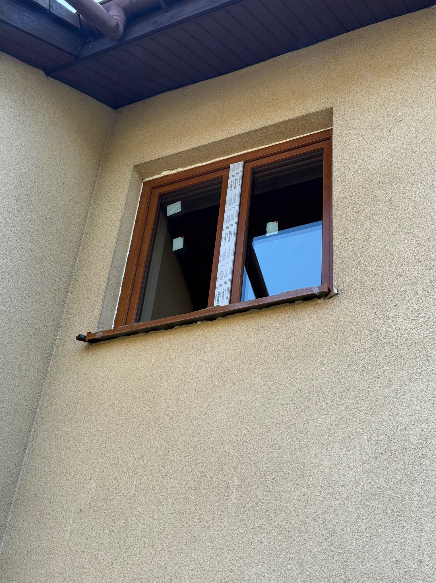 Brązowe okno z folią ochronną na ramie, zamontowane w elewacji budynku o beżowej strukturze. Widoczny fragment dachu. Ujęcie z dołu.