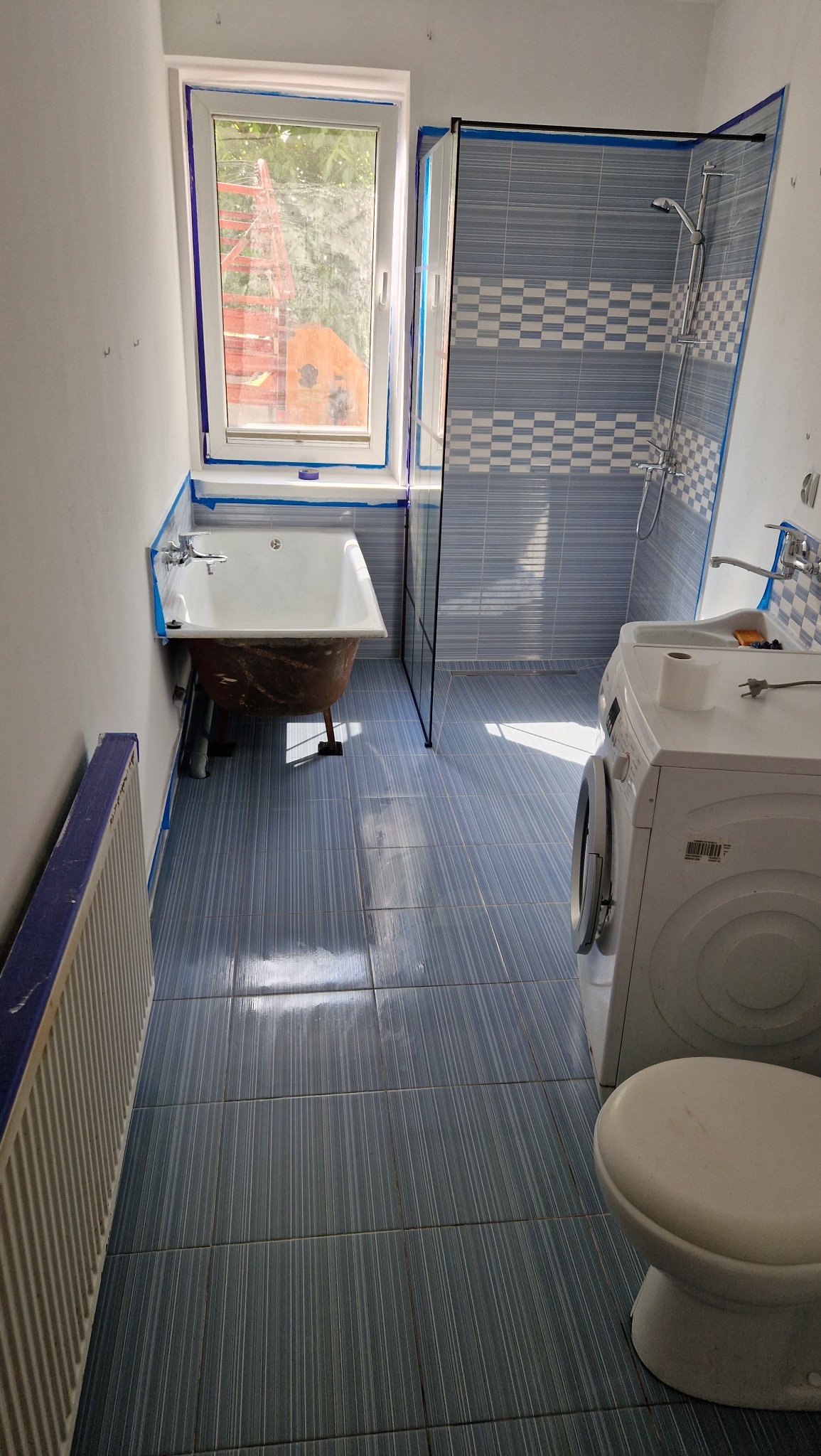 Niebieska łazienka po remoncie z wanną na nóżkach, kabiną prysznicową i pralką. Zabezpieczenia taśmą malarską na krawędziach. Widoczne płytki podłogowe i ścienne.