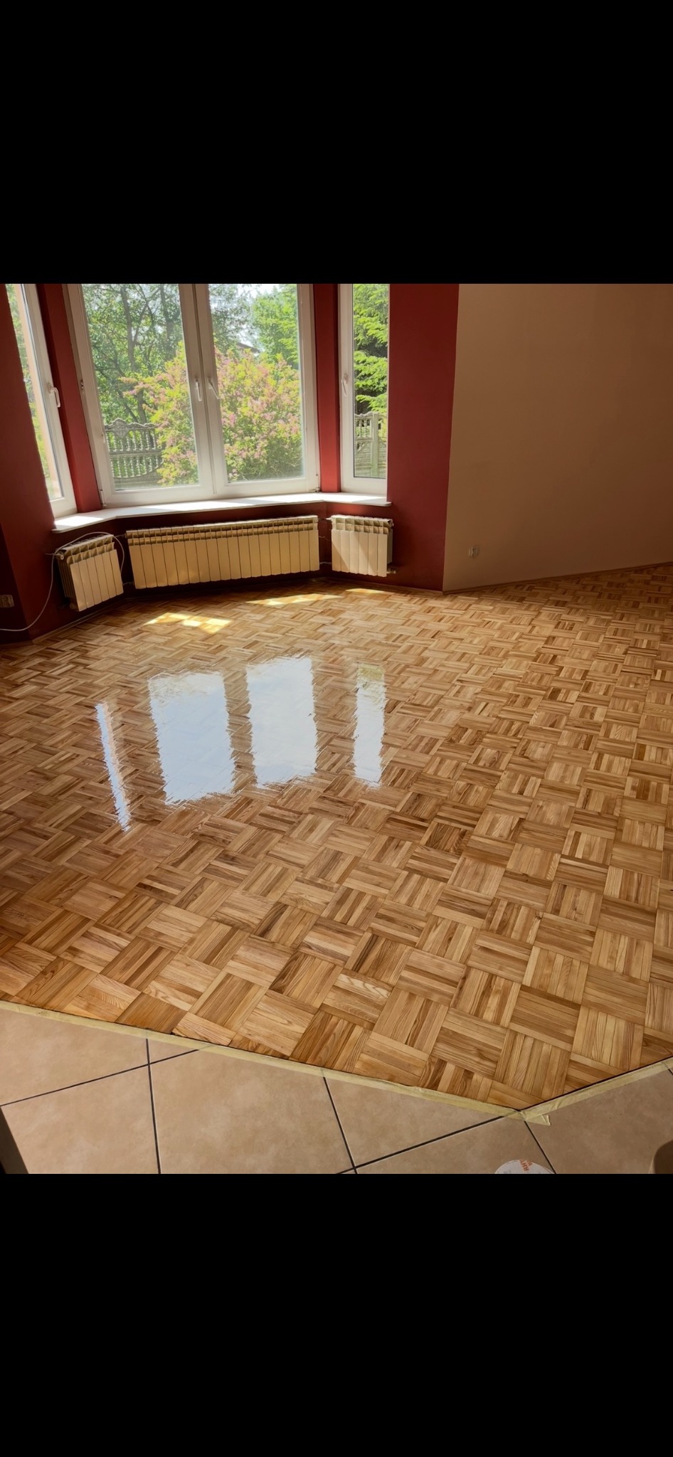 Błyszcząca, odnowiona podłoga parkietowa w jodełkę, po renowacji. Widoczne okno z widokiem na zieleń i trzy grzejniki. Ściany w kolorze bordo i beżu.