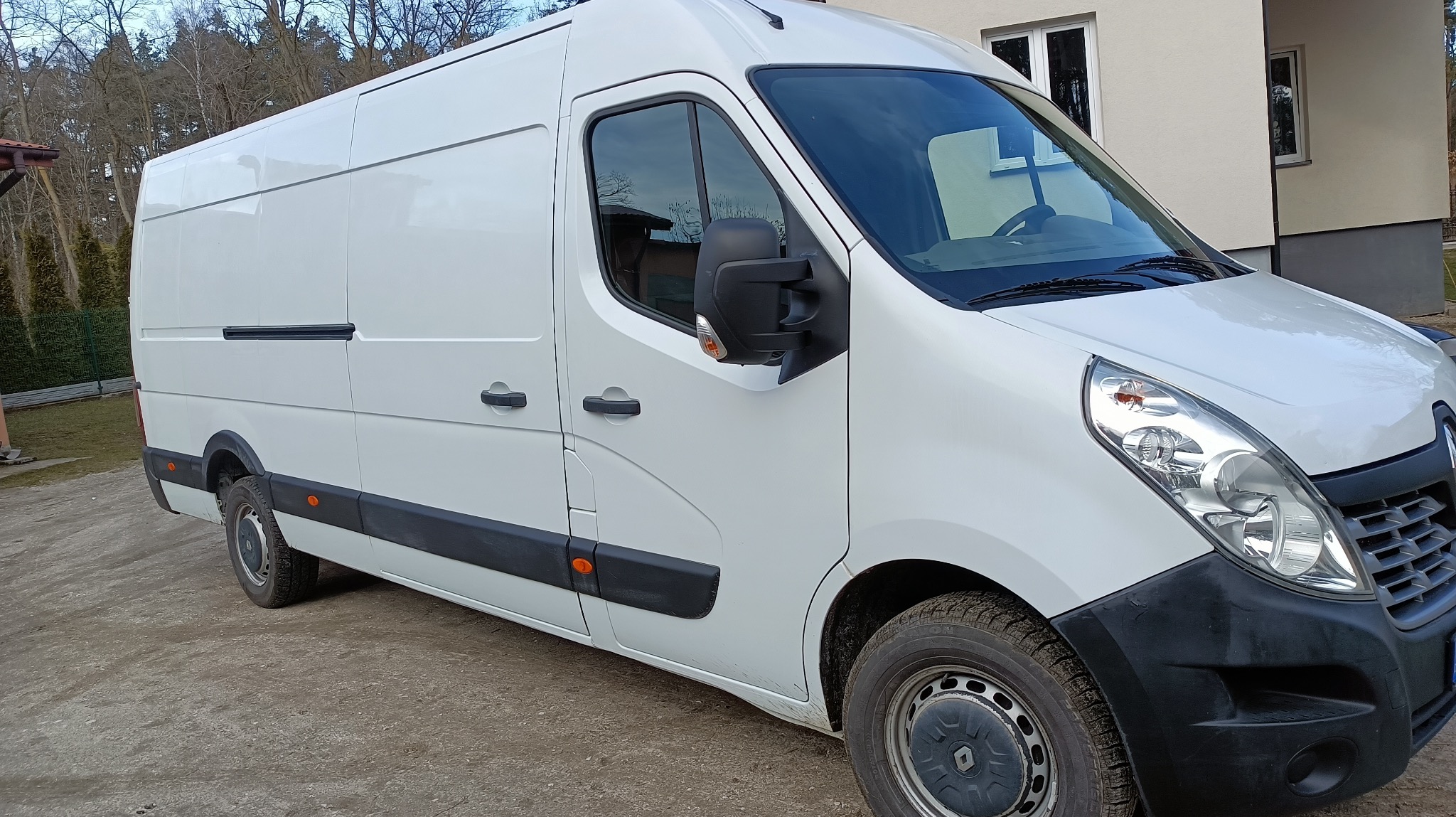 Biały Renault Master, widok z boku, zaparkowany na żwirowym podłożu przed budynkiem. Widoczne detale: relingi dachowe, lusterka, opony. Dzień, naturalne oświetlenie.