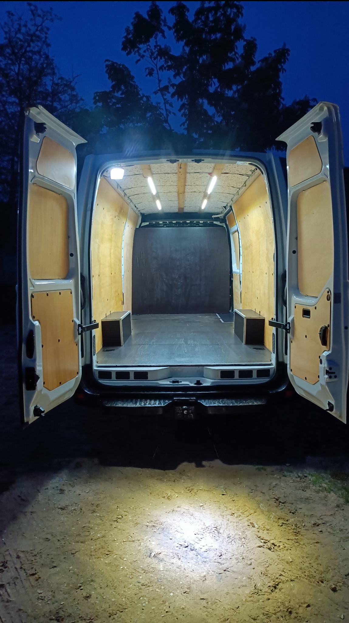 Wnętrze dostawczego busa z otwartymi drzwiami, wyłożone sklejką i oświetlone ledami. Widoczne dwa małe schowki. Zewnętrzne oświetlenie pada na ziemię.