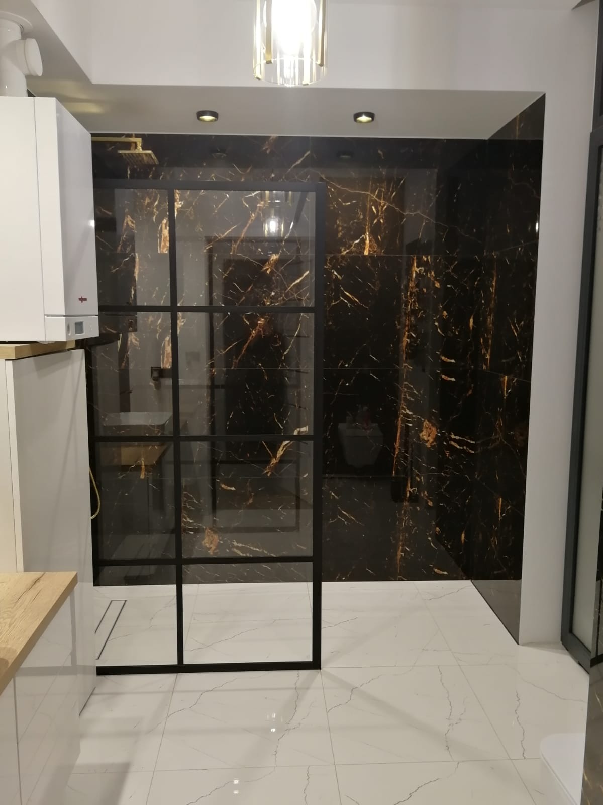 Nowoczesna łazienka z czarnymi płytkami imitującymi marmur ze złotymi żyłami, kabiną prysznicową typu walk-in z czarnymi szprosami i białymi płytkami podłogowymi.
