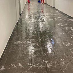CzystoSPACE - Mycie podłogi z płytek w korytarzu. Widoczne ślady po mopie i resztki piany. W tle osoby w czerwonych kombinezonach i wózek do sprzątania.