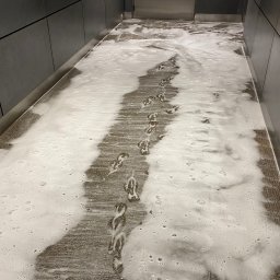 CzystoSPACE - Dywan pokryty pianą czyszczącą z widocznymi śladami stóp, sugerujący etap prania. Szare płytki na ścianach i metalowe wykończenia dodają nowoczesnego charakteru wnętrzu.