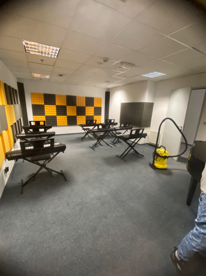 Sala muzyczna z pianinami Yamaha i żółtym odkurzaczem. Akustyczne panele na ścianach. Szara wykładzina. Widok na salę podczas sprzątania.