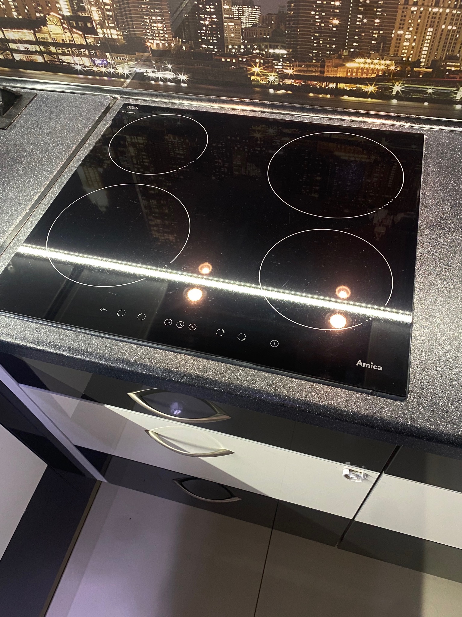 Czarna płyta indukcyjna Amica z czterema polami grzewczymi, zamontowana w blacie kuchennym na tle tapety z panoramą miasta nocą. Widoczne szafki kuchenne z białymi frontami.