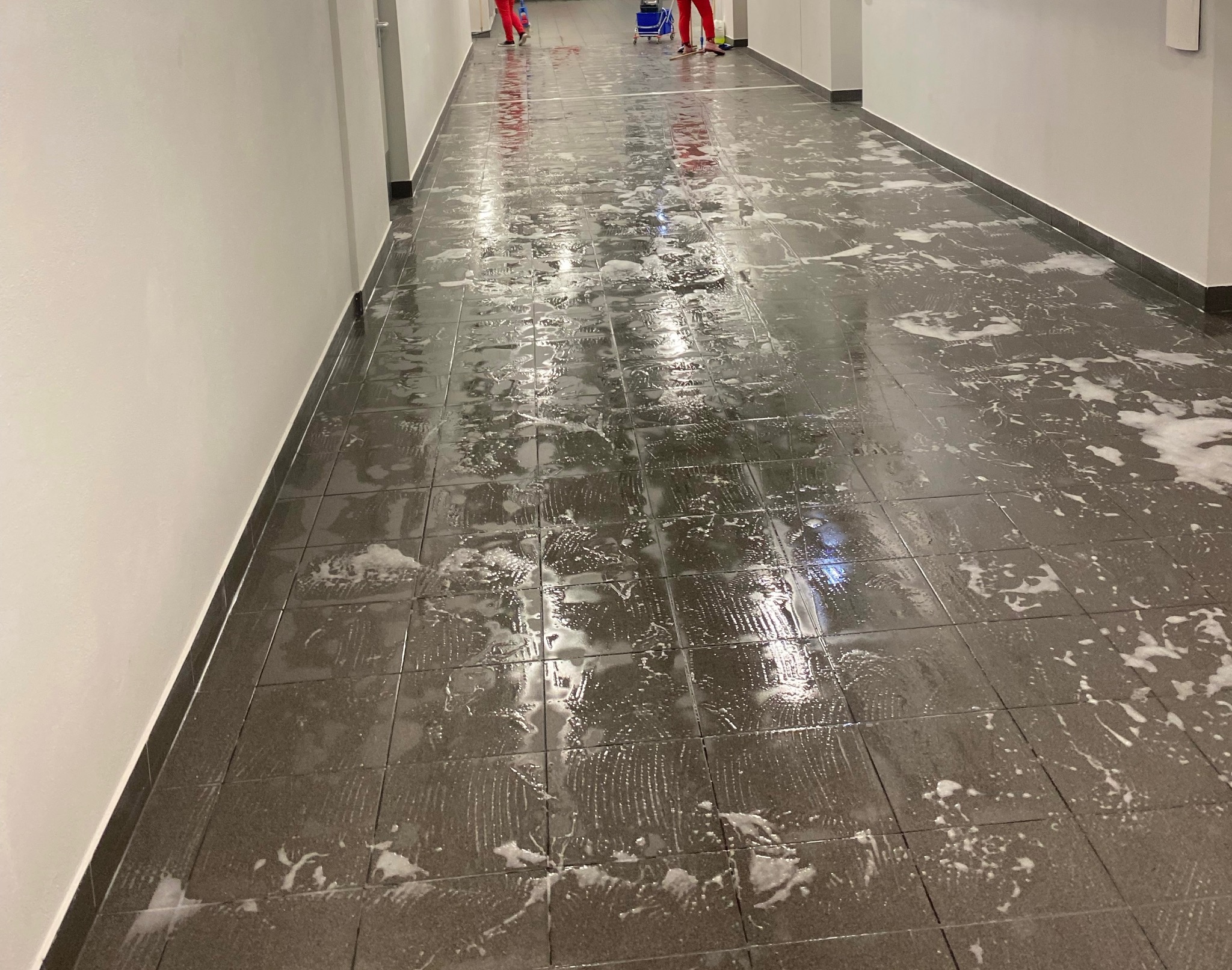 Mycie podłogi z płytek w korytarzu. Widoczne ślady po mopie i resztki piany. W tle osoby w czerwonych kombinezonach i wózek do sprzątania.
