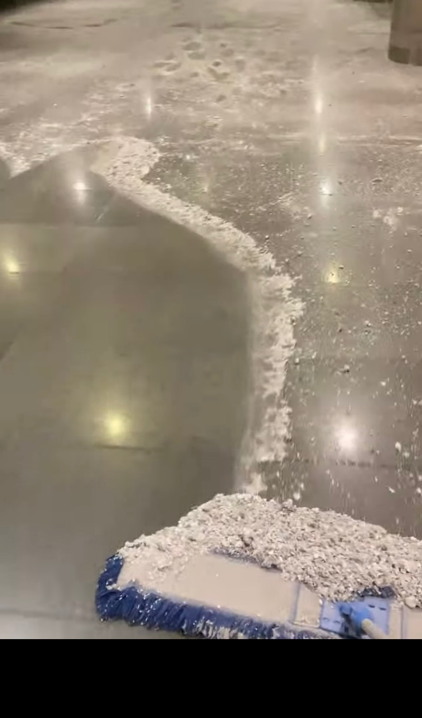 Mop z niebieskim włosiem zbiera biały pył z polerowanej podłogi. Widoczne ślady po czyszczeniu. Oświetlenie sufitowe odbija się w podłodze.