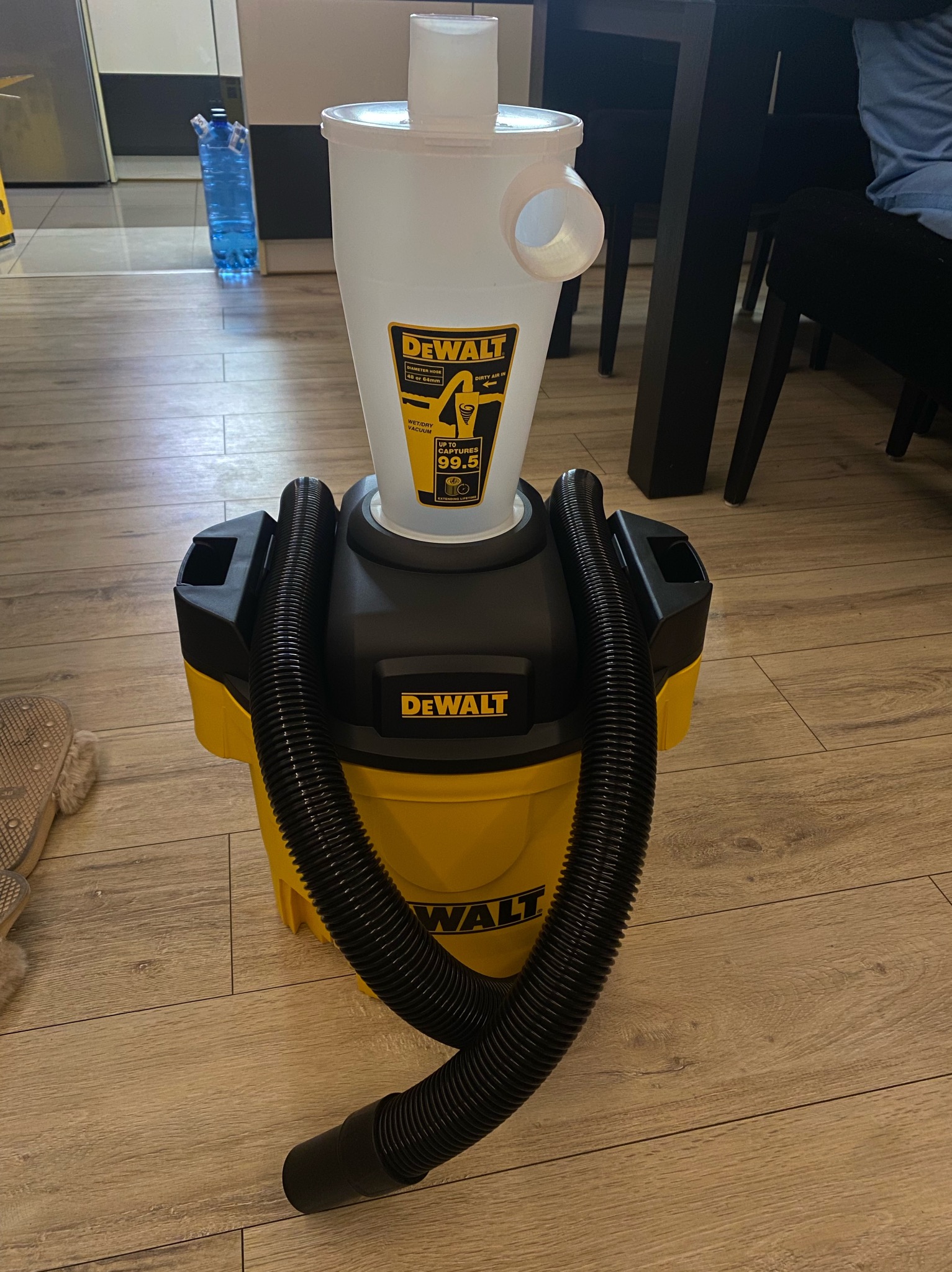 Żółto-czarny odkurzacz przemysłowy DeWalt z separatorem cyklonowym na drewnianej podłodze. Widoczny wąż ssący i pojemnik na zanieczyszczenia. W tle butelka z wodą.