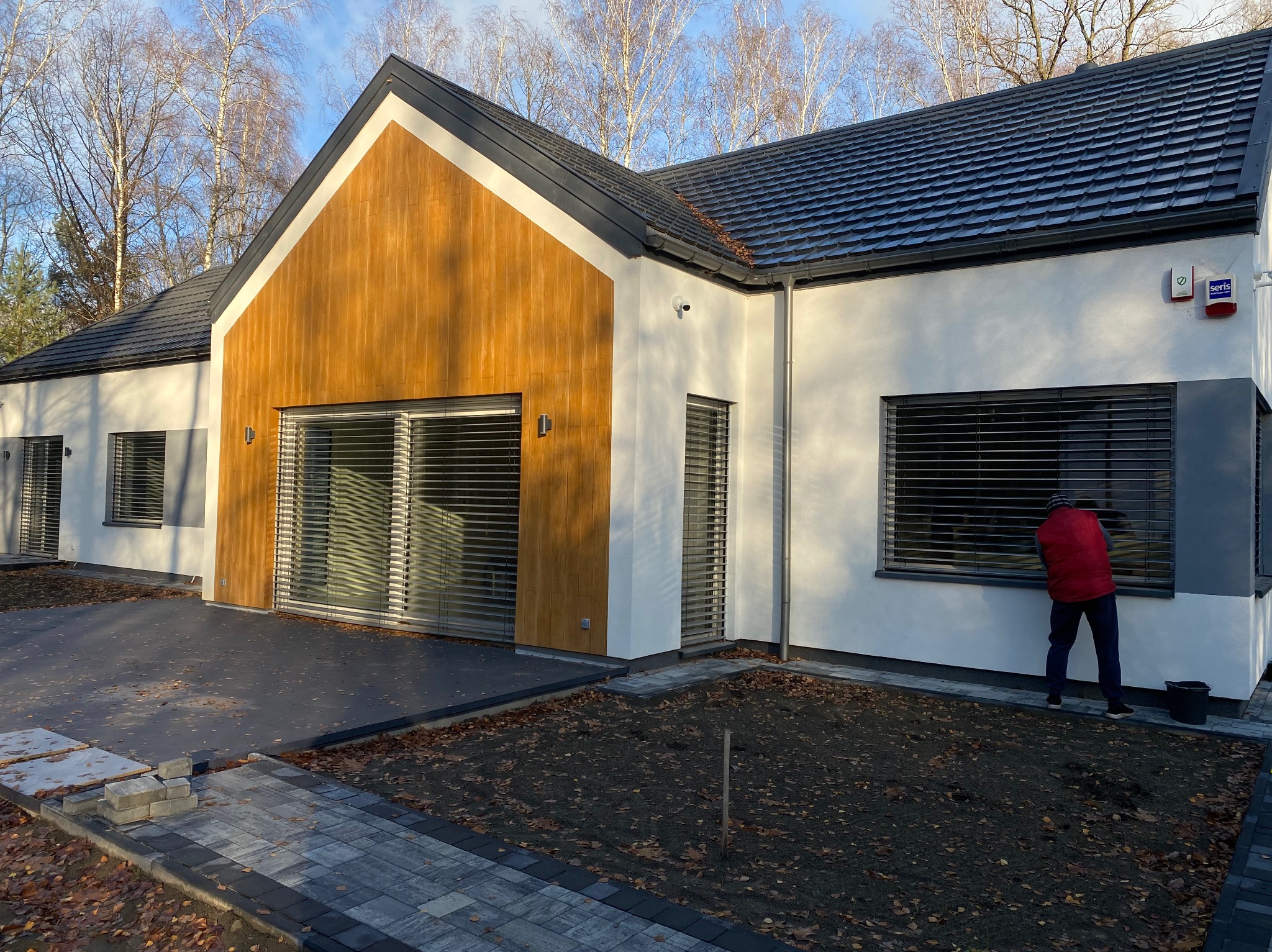 Osoba w czerwonej kamizelce myje okna nowoczesnego domu z drewnianą fasadą. Widoczne ułożone kostki brukowe i przygotowany teren pod trawnik.