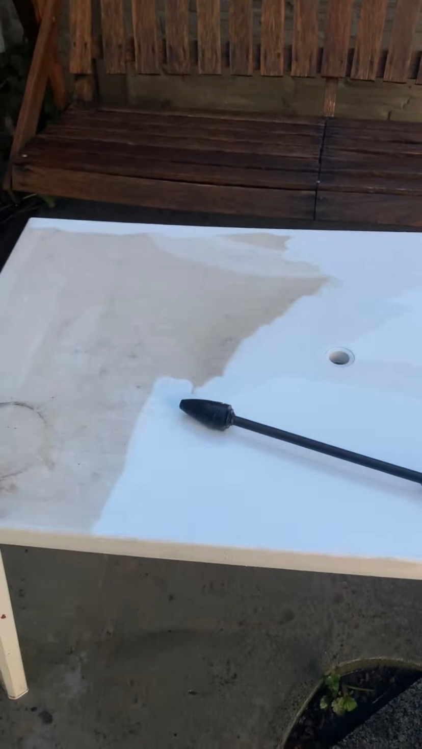 Czyszczenie białego stołu ogrodowego myjką ciśnieniową. Widoczna różnica między czystą a zabrudzoną powierzchnią. W tle drewniana ławka.