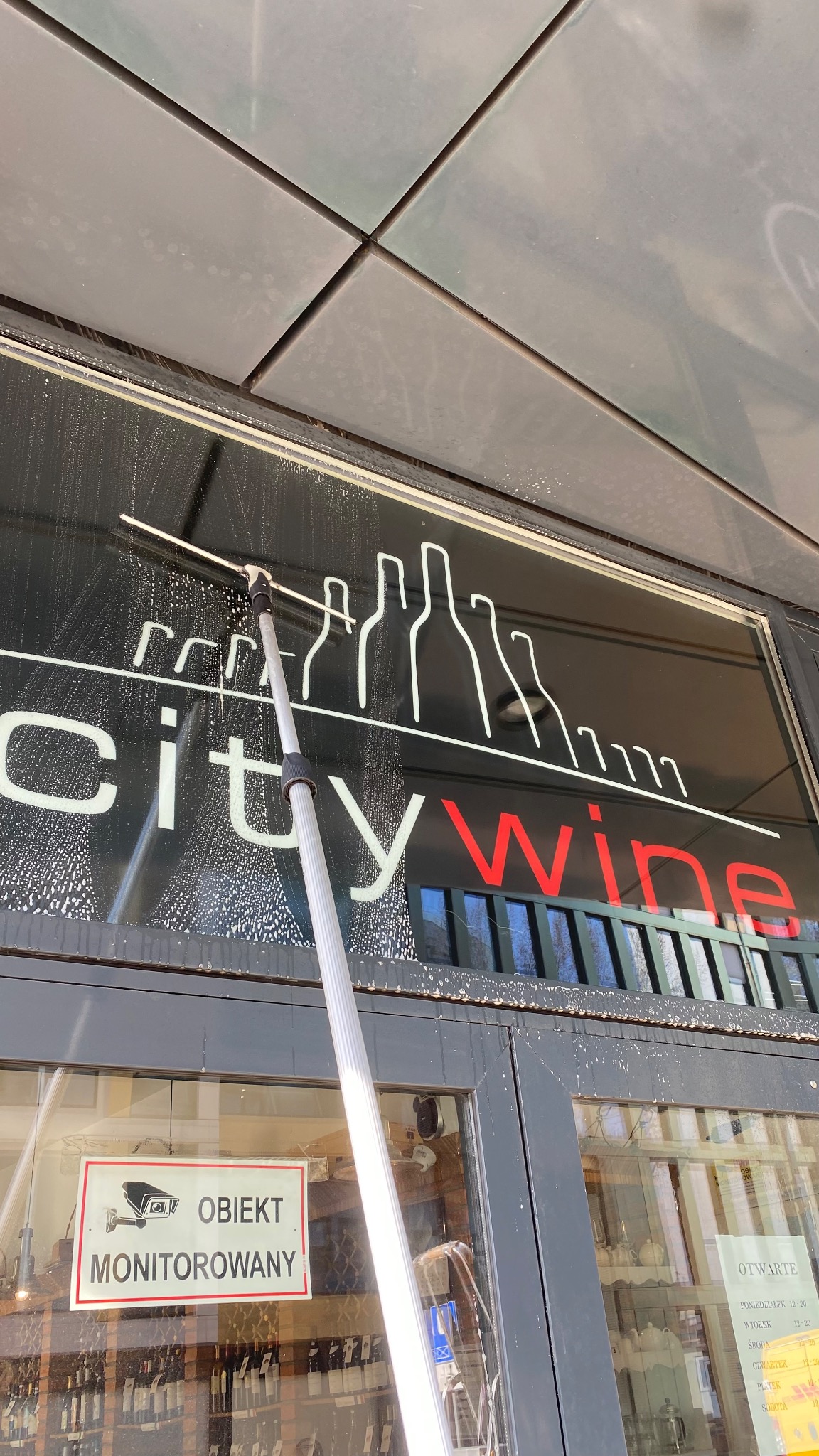 Mycie okna wystawowego z grafiką 'City Wine' za pomocą mopa teleskopowego. Widoczne ślady detergentu i odbicie wnętrza sklepu z winami. Ujęcie z dołu.