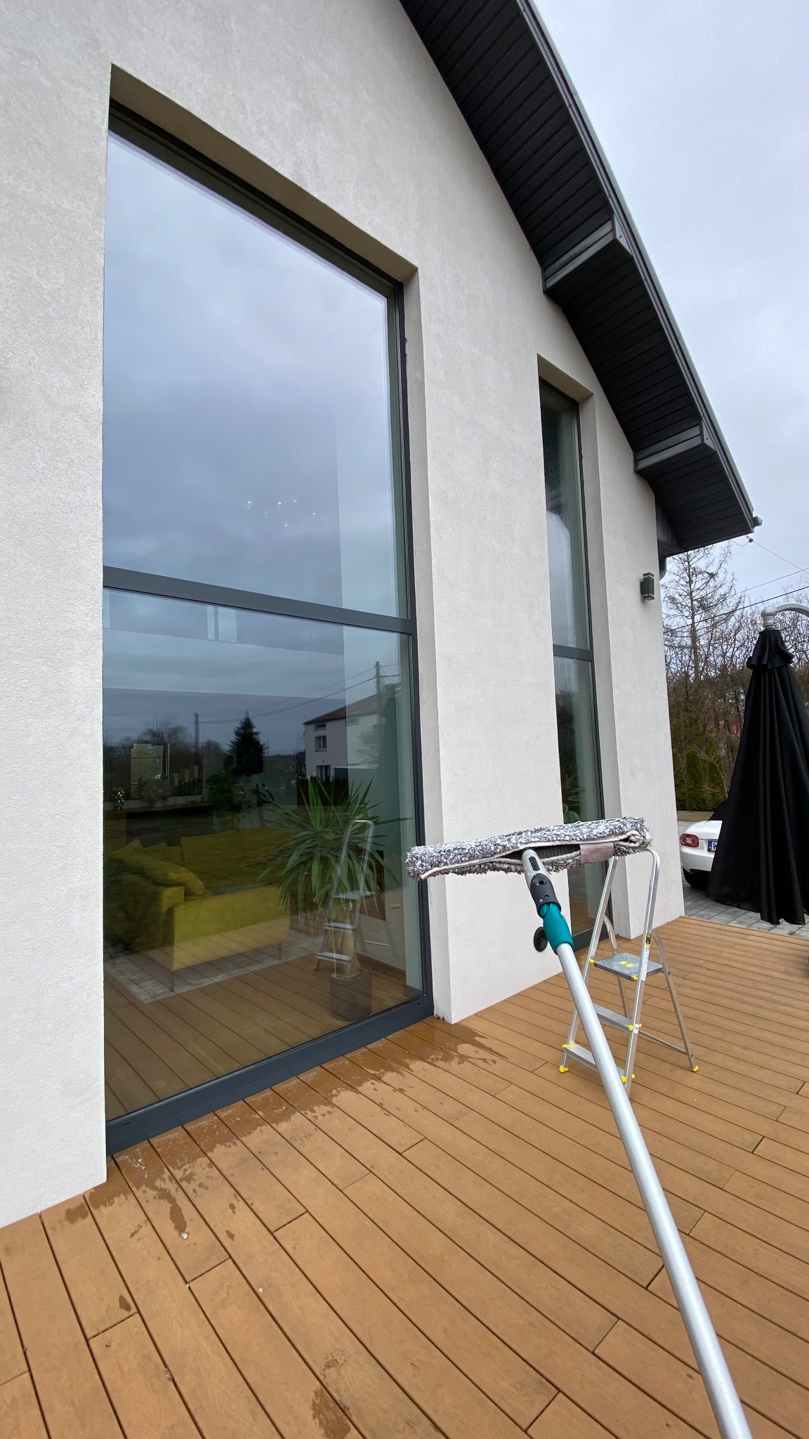 Mycie okien domu jednorodzinnego z użyciem profesjonalnego sprzętu. Na pierwszym planie kij teleskopowy z myjką, w tle drabina i duże okno z widokiem na wnętrze.